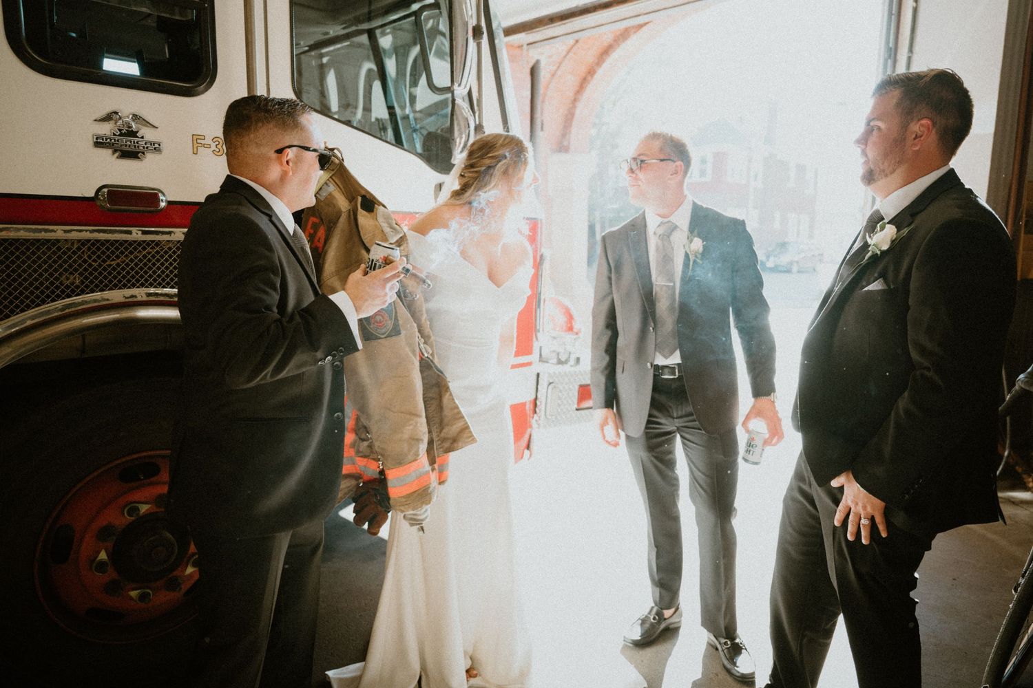 Bride and groom celebrate with wedding guests beside a fire truck inside a bright firehouse.