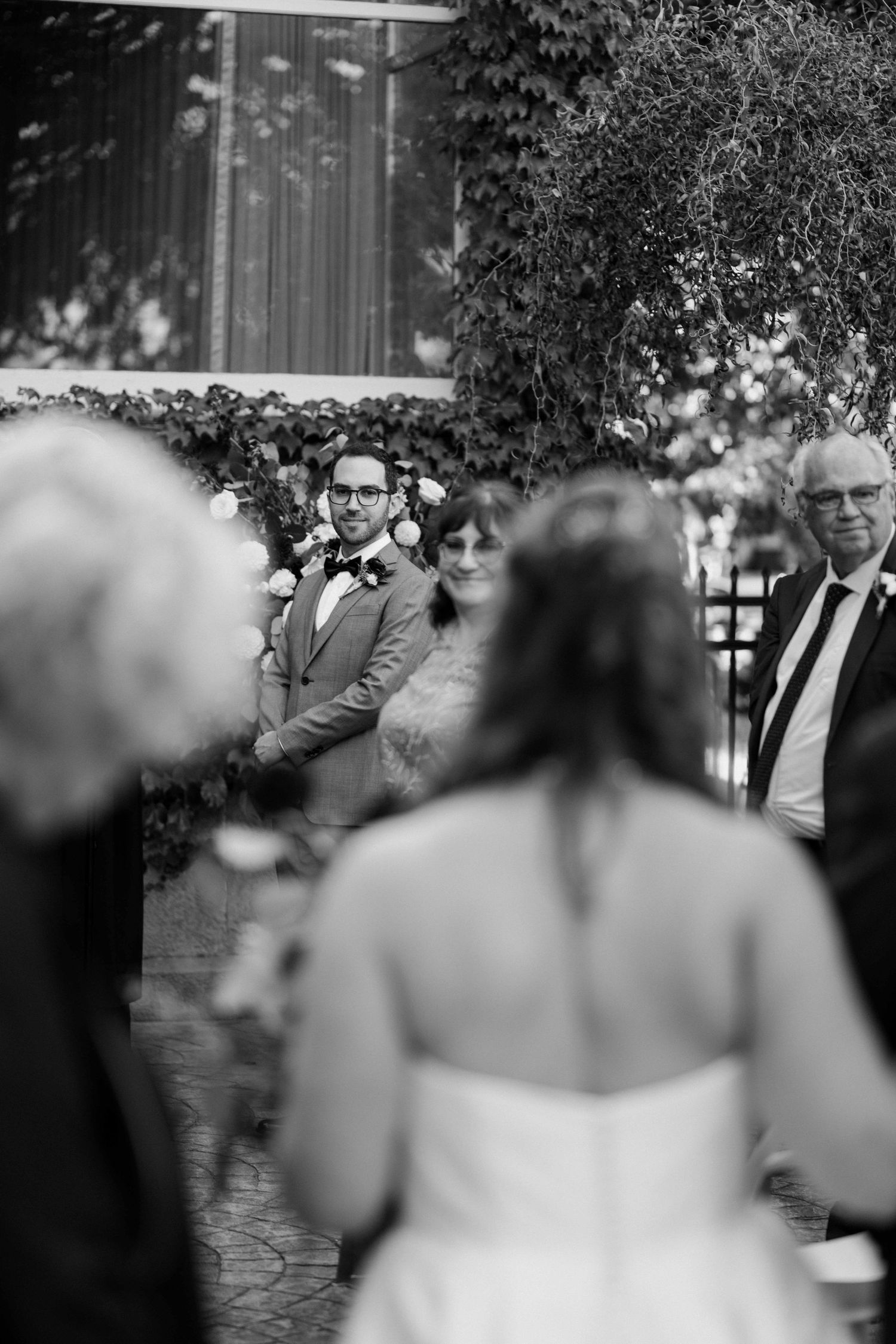 Black and white photo of groom watching bride walk down the aisle at outdoor wedding ceremony.