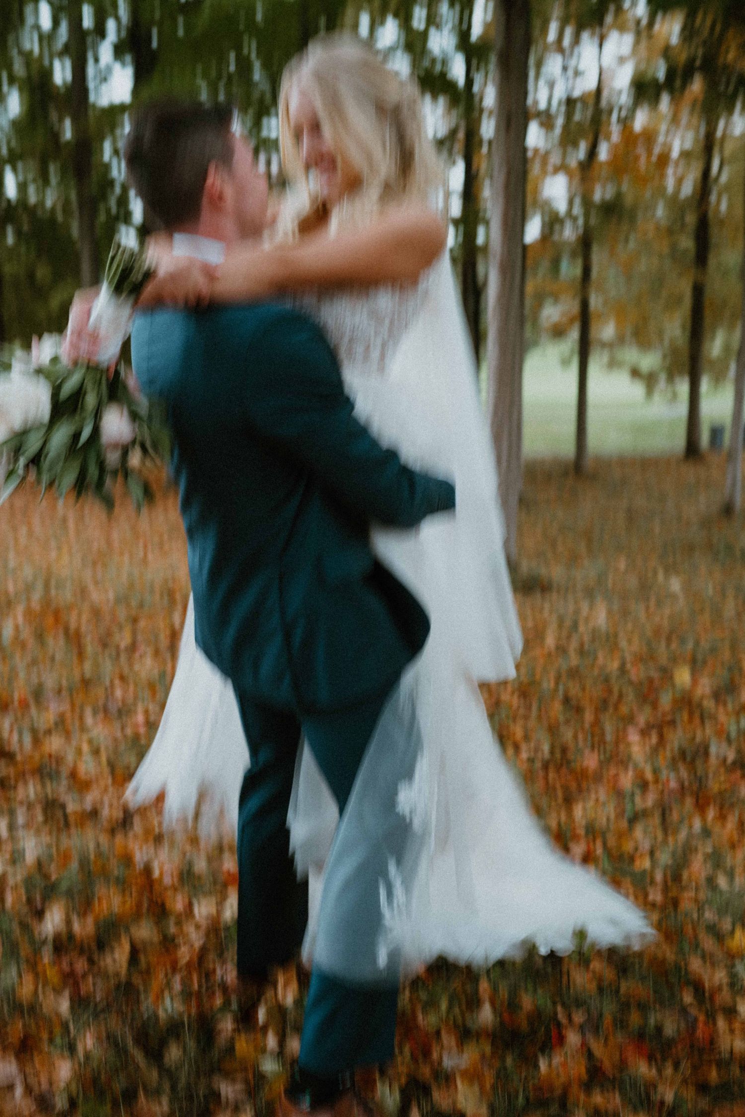 Blurred artistic shot of groom lifting bride in white dress on autumn leaves.