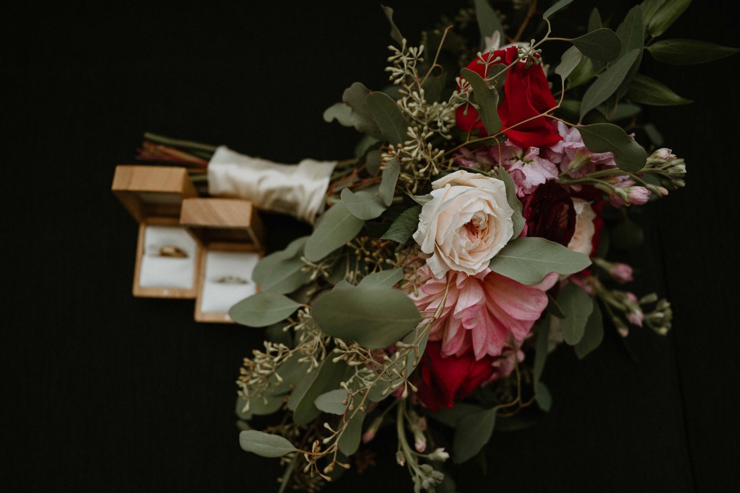 Bridal bouquet with red roses, pink peonies, and eucalyptus beside gold ring boxes.