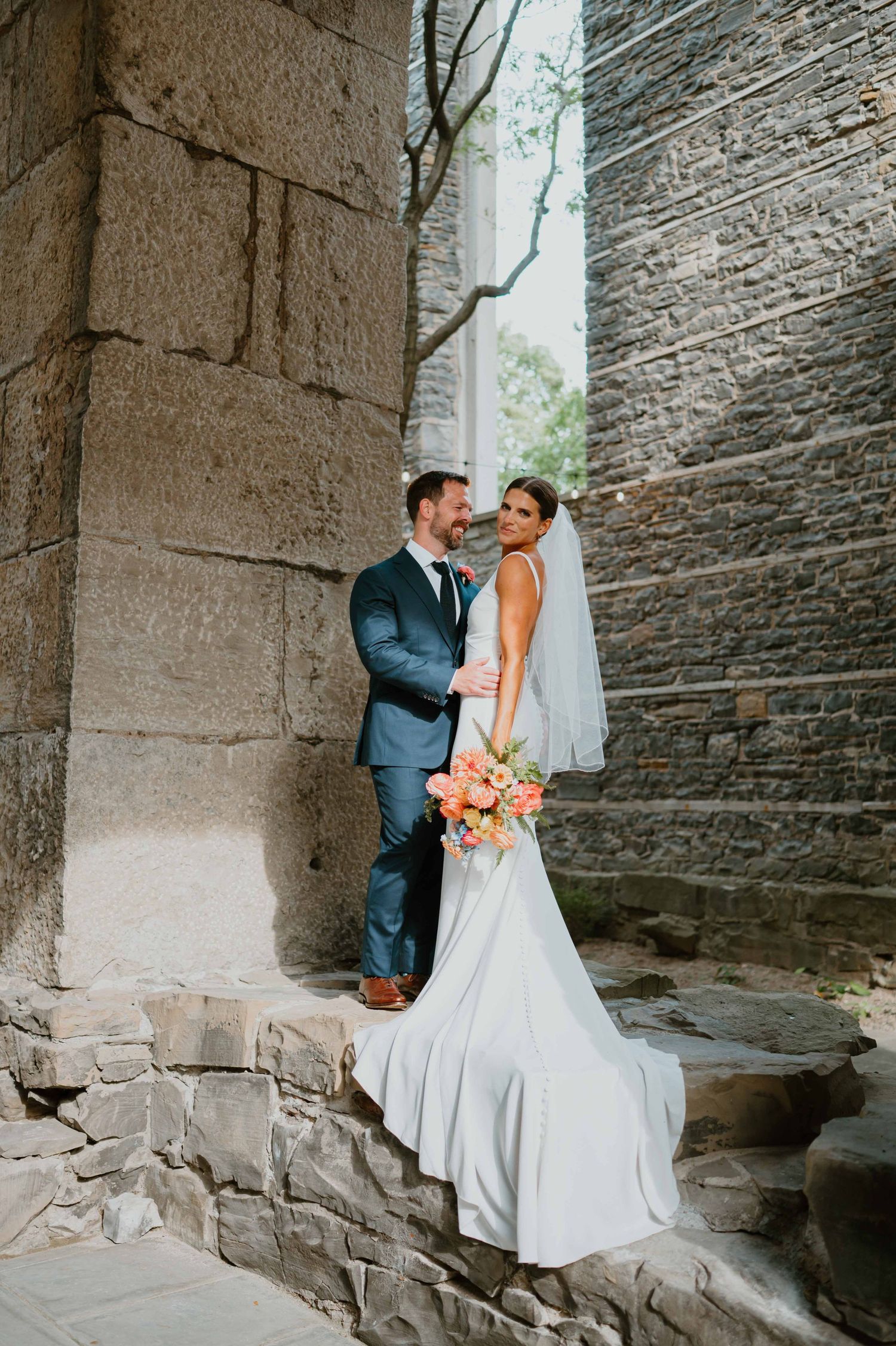 Bride holding coral bouquet and groom in navy suit pose romantically between stone pillars at historic wedding venue.