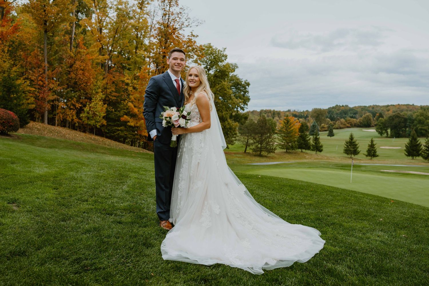 Bride and groom posing on golf course with autumn foliage background.