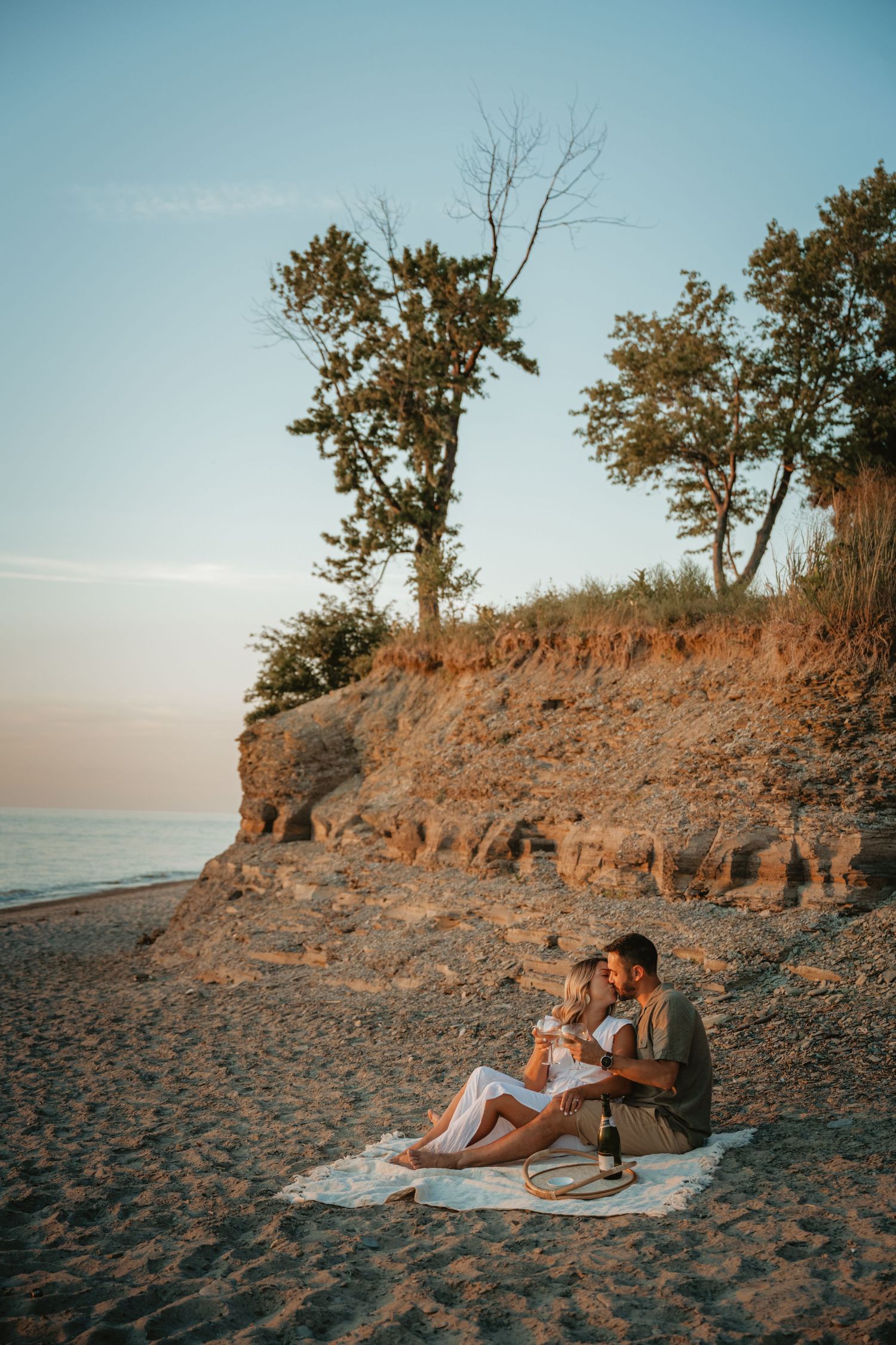 Couple sits together on a blanket for a romantic beach picnic at golden hour near a sandy cliff.