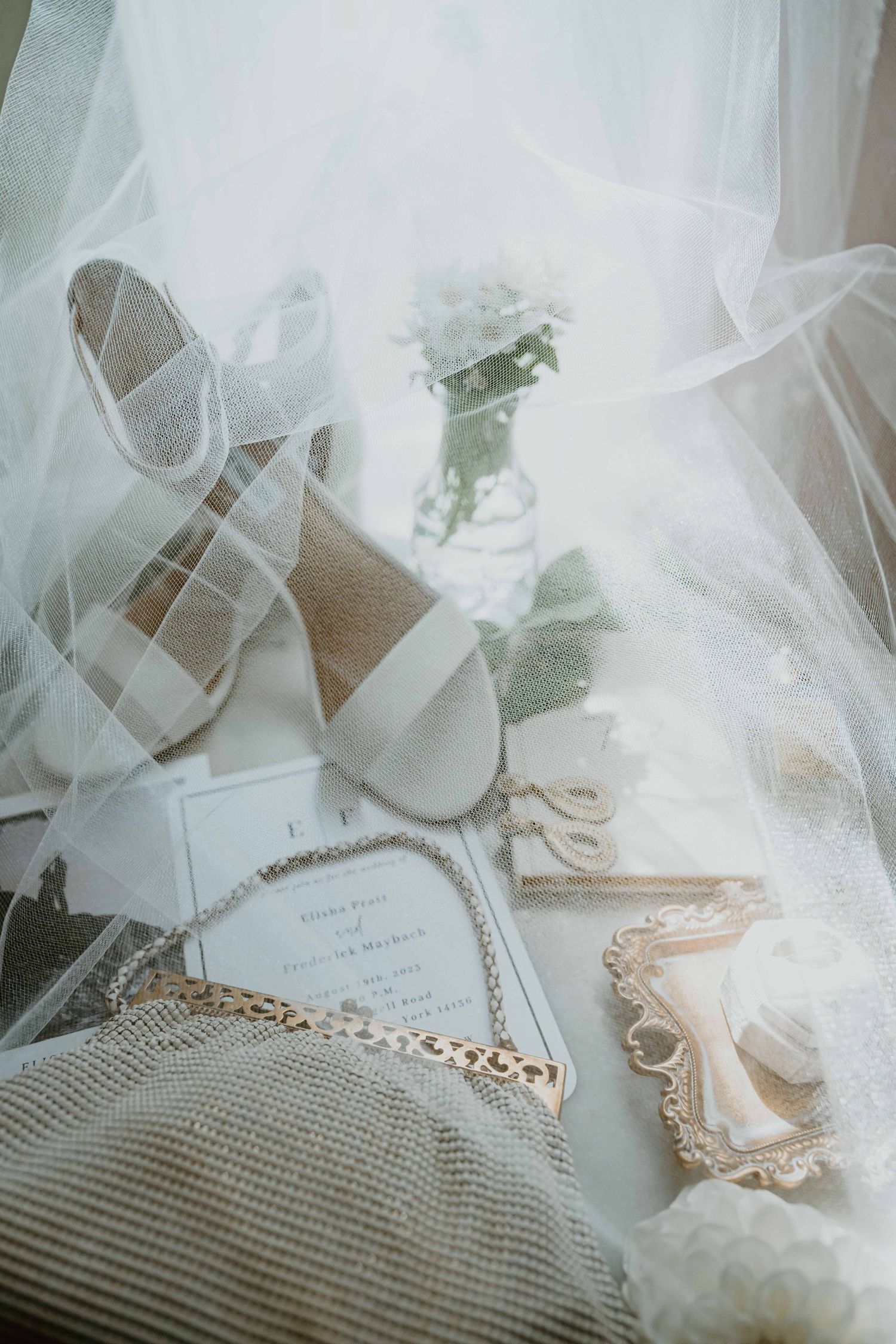 Elegant bridal flat lay with lace heels, gold jewelry, pearl necklace, and wedding invitation draped in tulle veil.