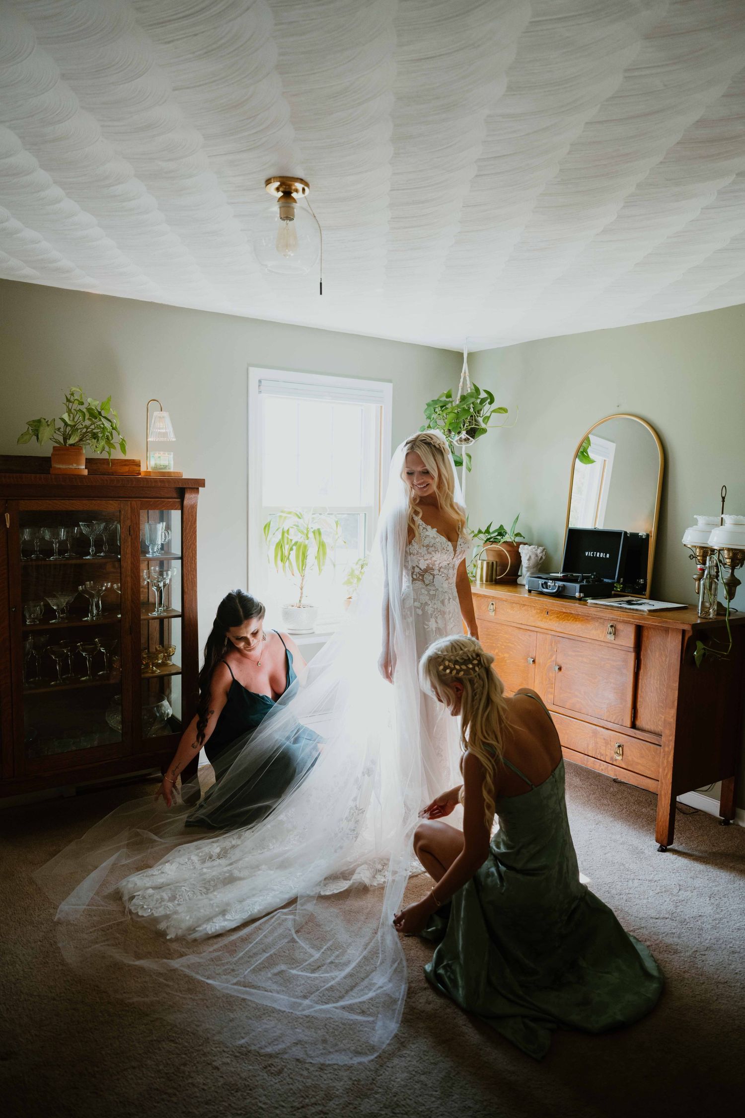 Bridesmaids kneeling to spread bride's lace wedding gown train in warmly decorated bedroom.