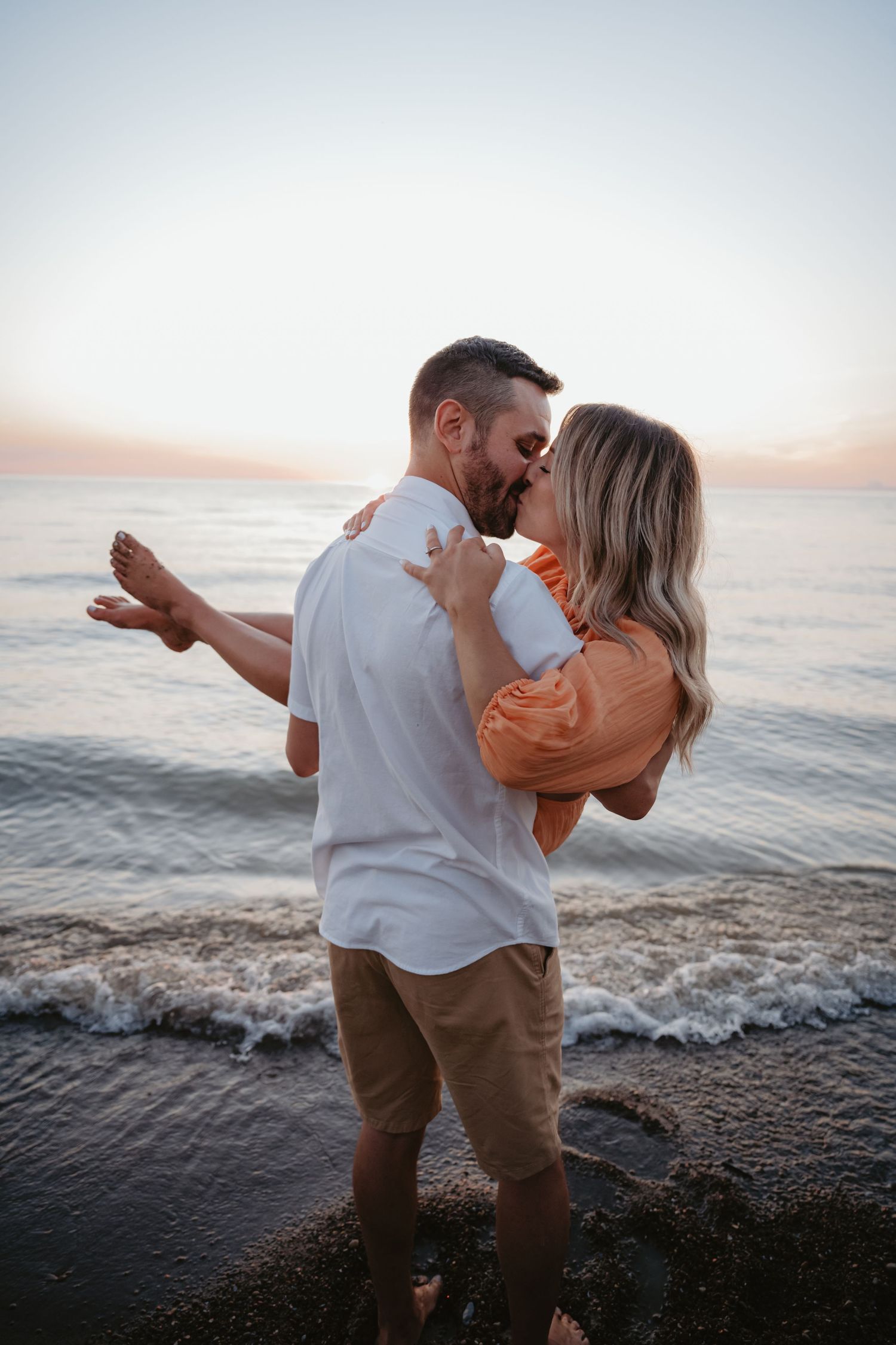 Couple shares a romantic kiss at sunset on a rocky beach, man lifting woman in orange dress.