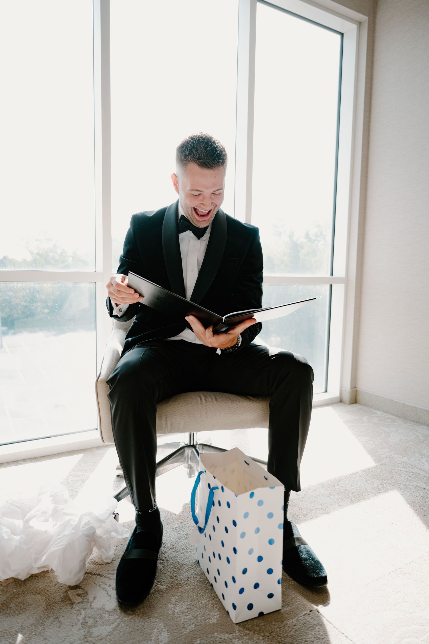 Groom in black tuxedo laughing while reading a book, seated by bright window with gift bag.
