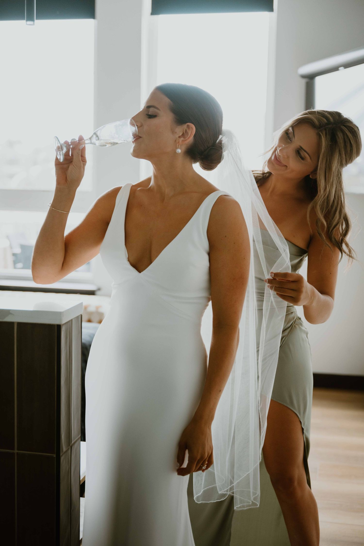 Bride in white gown sipping champagne while bridesmaid adjusts veil.