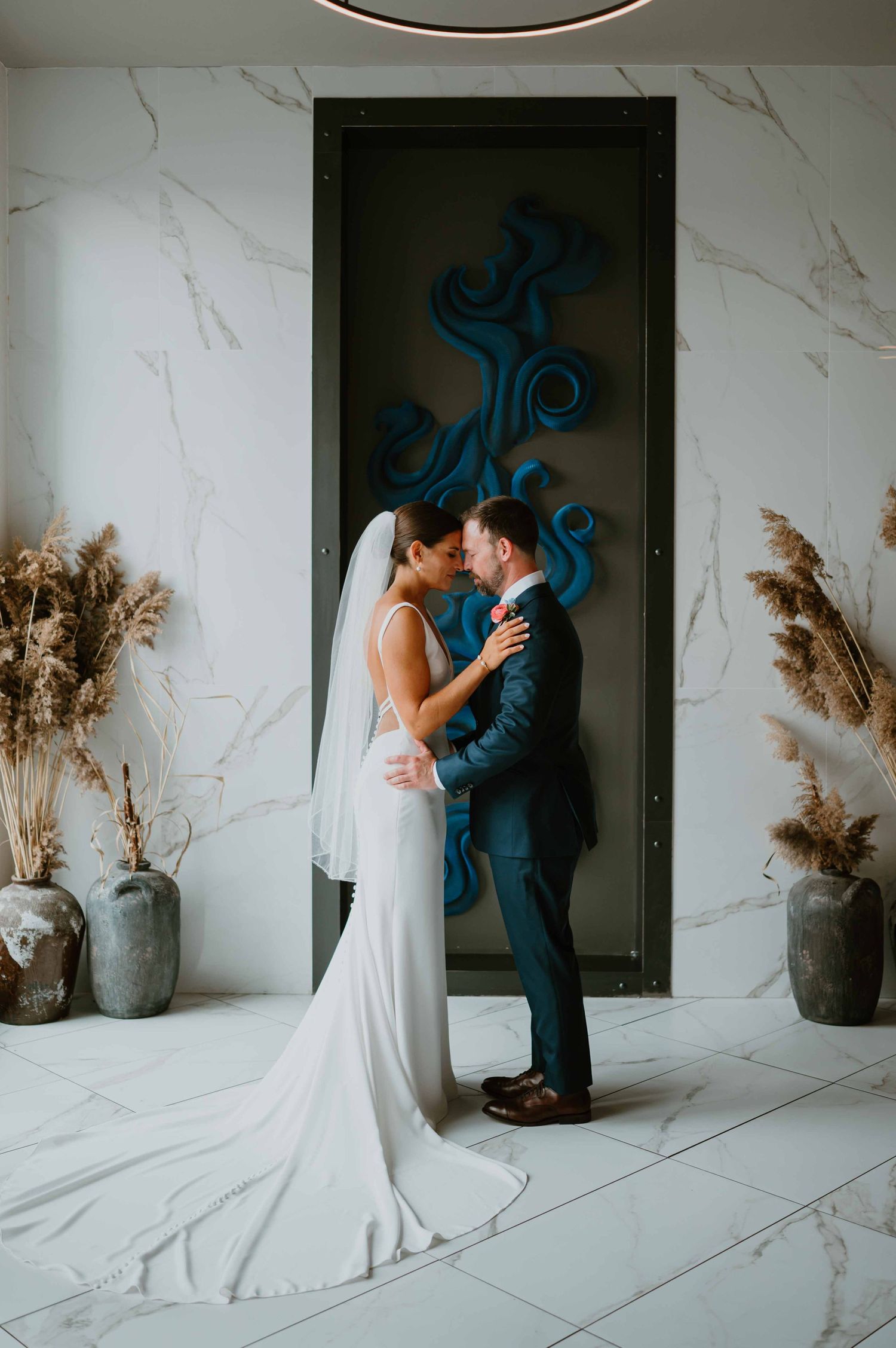 Bride and groom embrace in marble lobby with blue sculptural wall art.