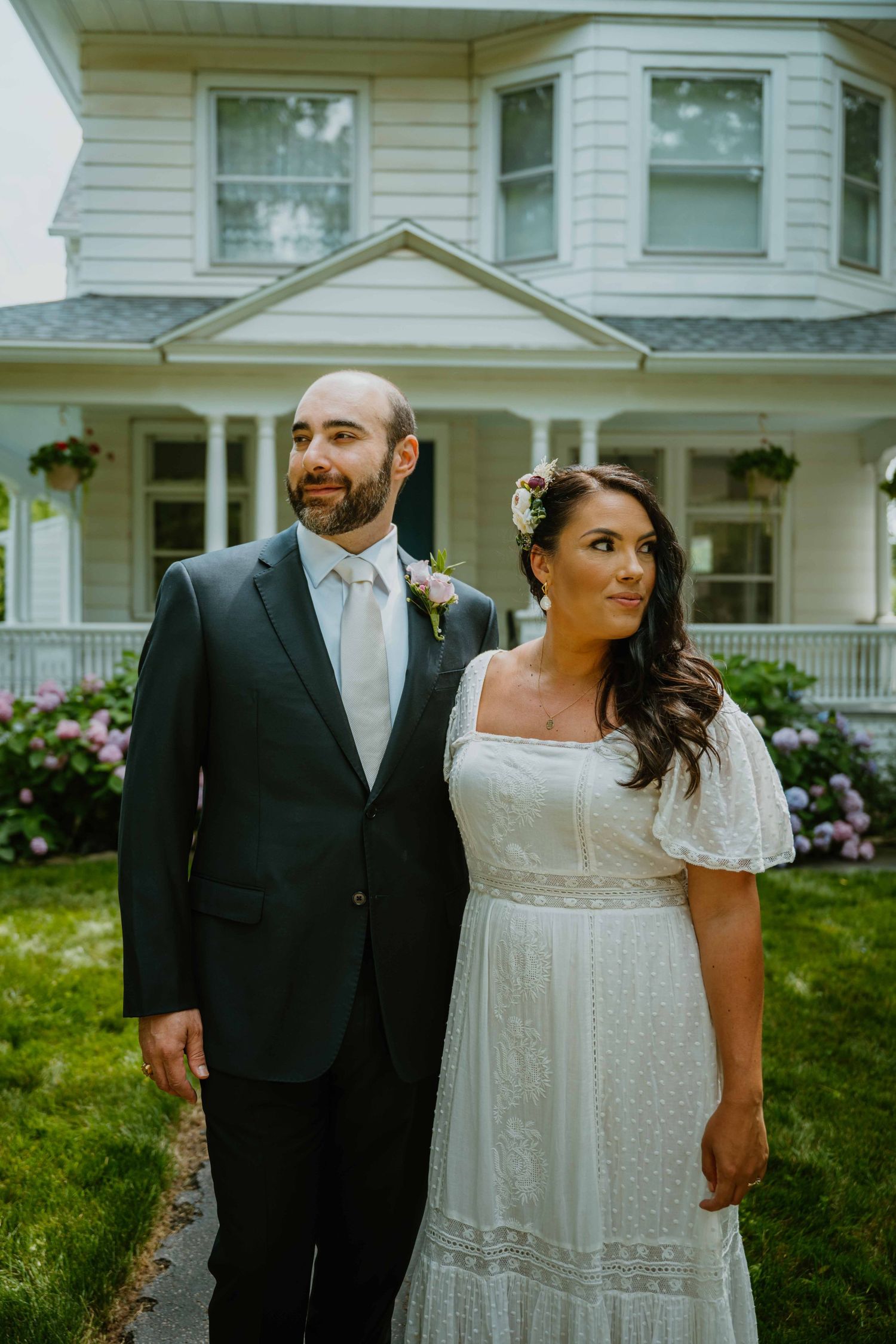 Bride and groom stand together in front of a white Victorian house with hydrangeas.