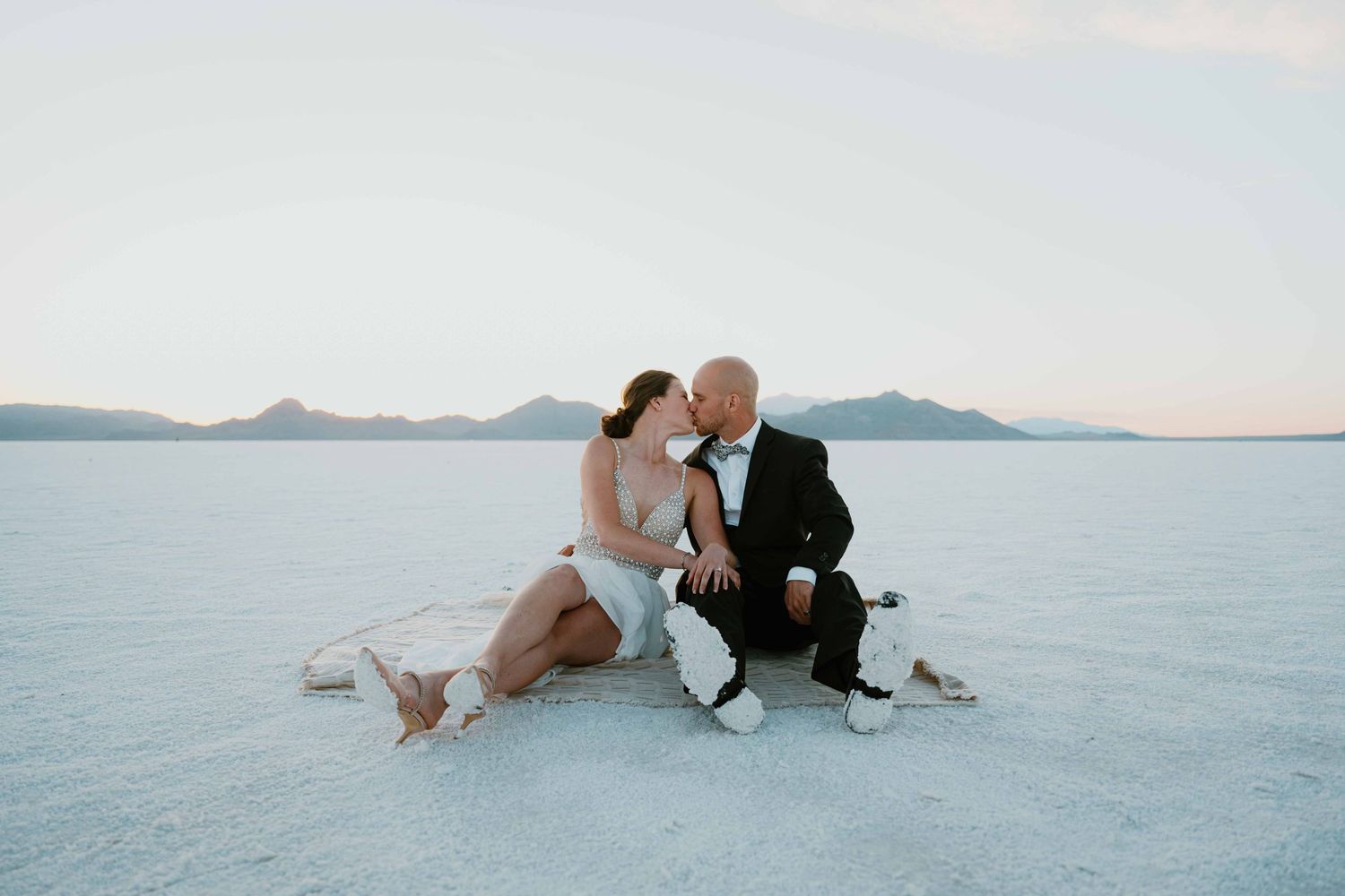 Couple shares a romantic kiss while sitting together on the Bonneville Salt Flats at dusk.