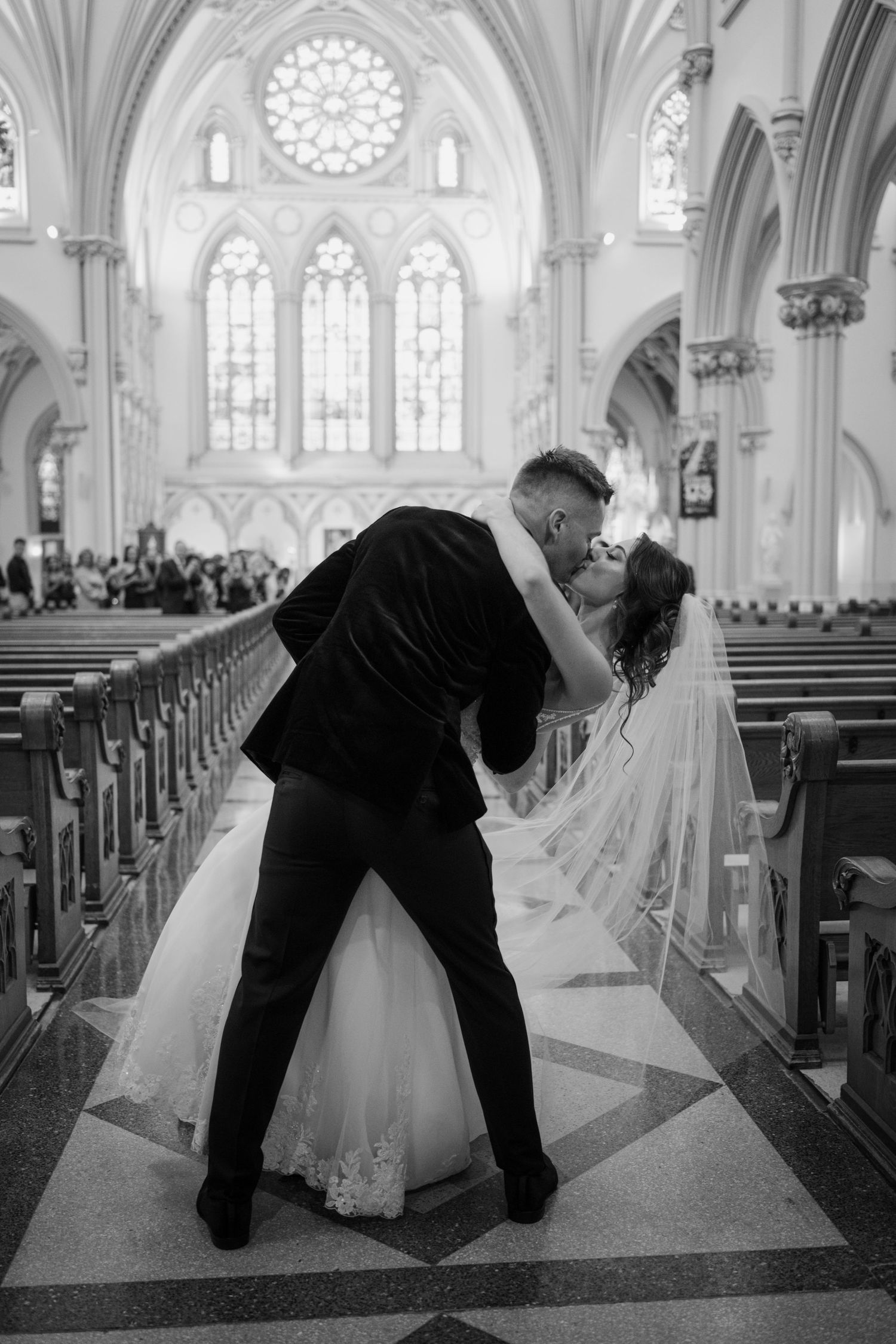 Black and white photo of groom dipping bride for romantic kiss in stunning Gothic cathedral aisle.