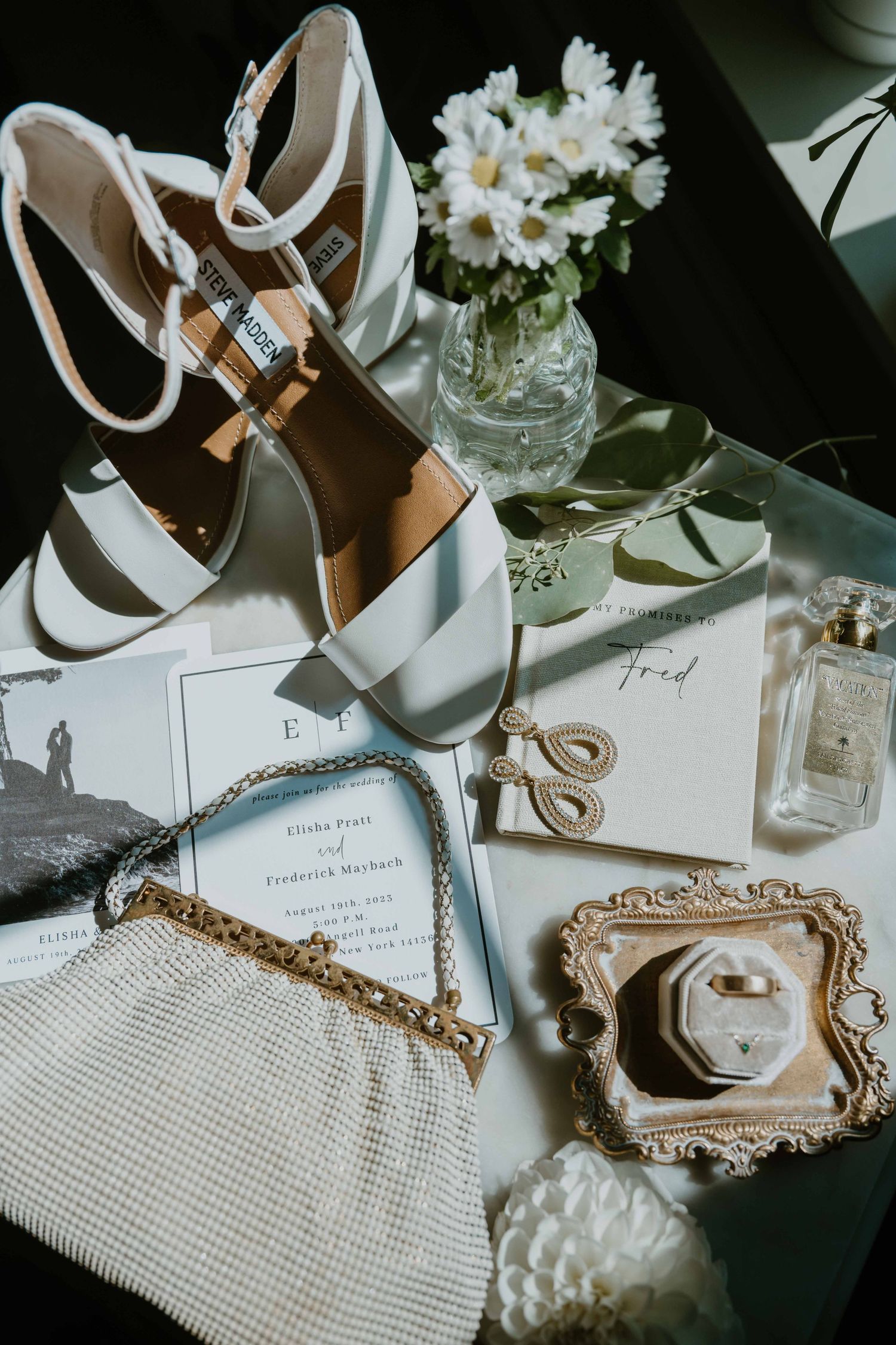 Flat lay of bridal accessories including white heels, gold jewelry, invitations, and perfume.