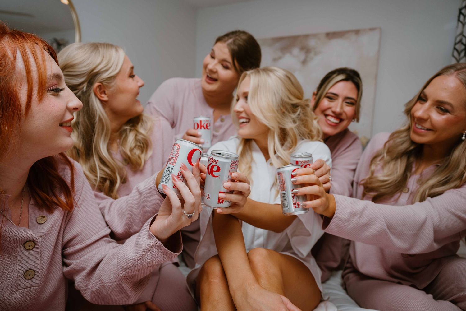 Bridesmaids in matching pink pajamas laughing and holding drinks together on a bed before the wedding.