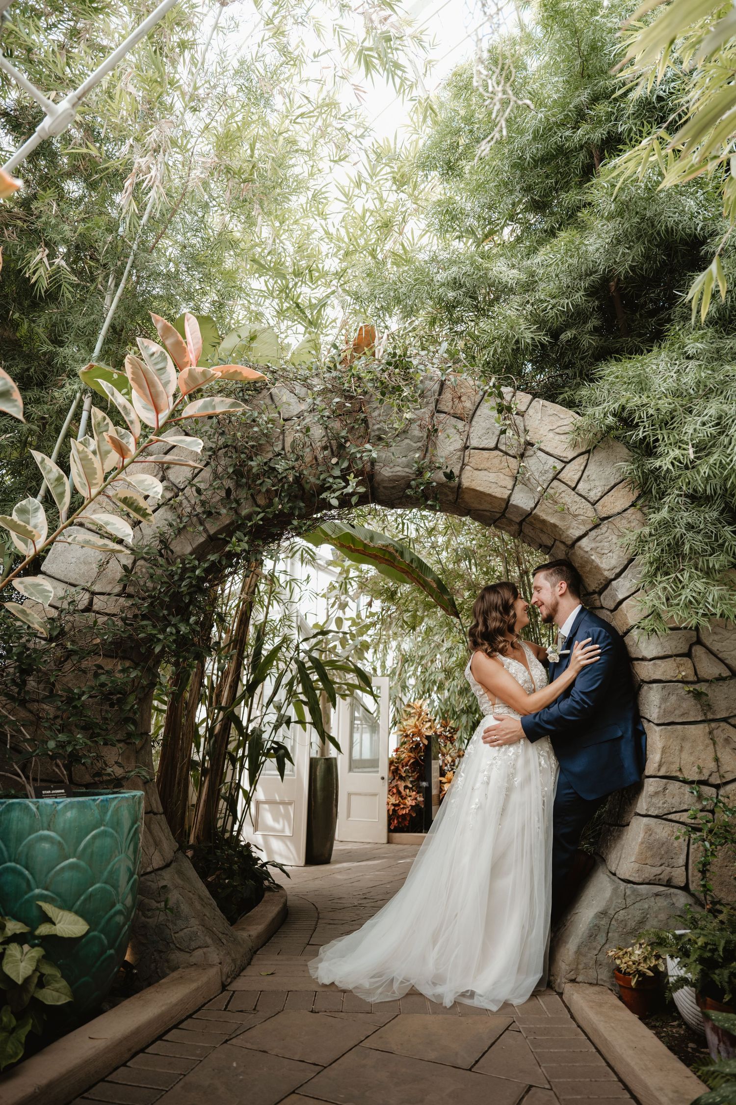 Wedding couple embracing under a rustic stone archway surrounded by lush tropical plants.