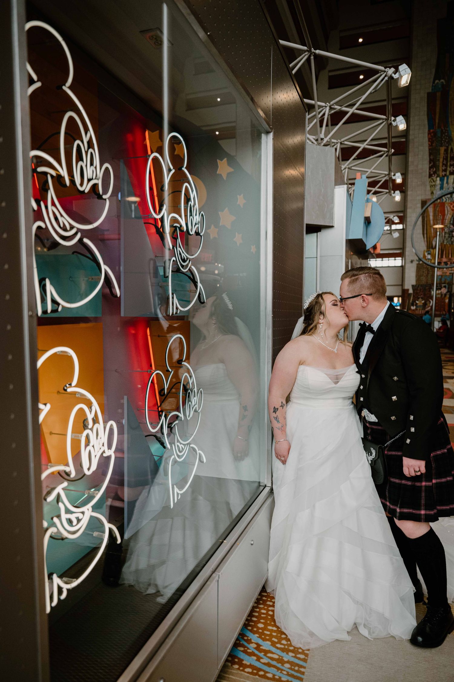Bride and groom share a kiss beside colorful Mickey Mouse neon art at Disney wedding.