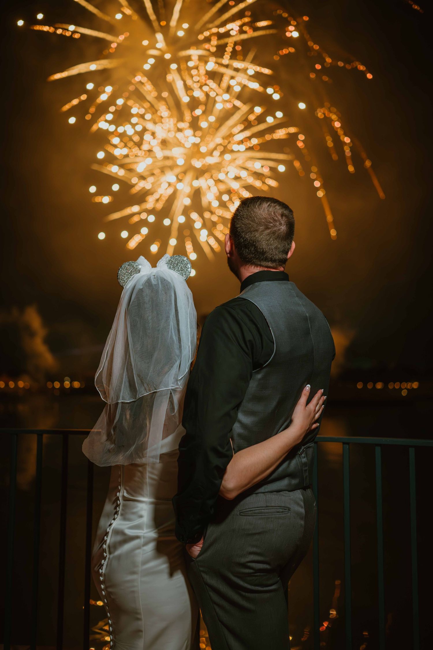 Bride and groom stand arm in arm watching golden fireworks explode over the water at night.