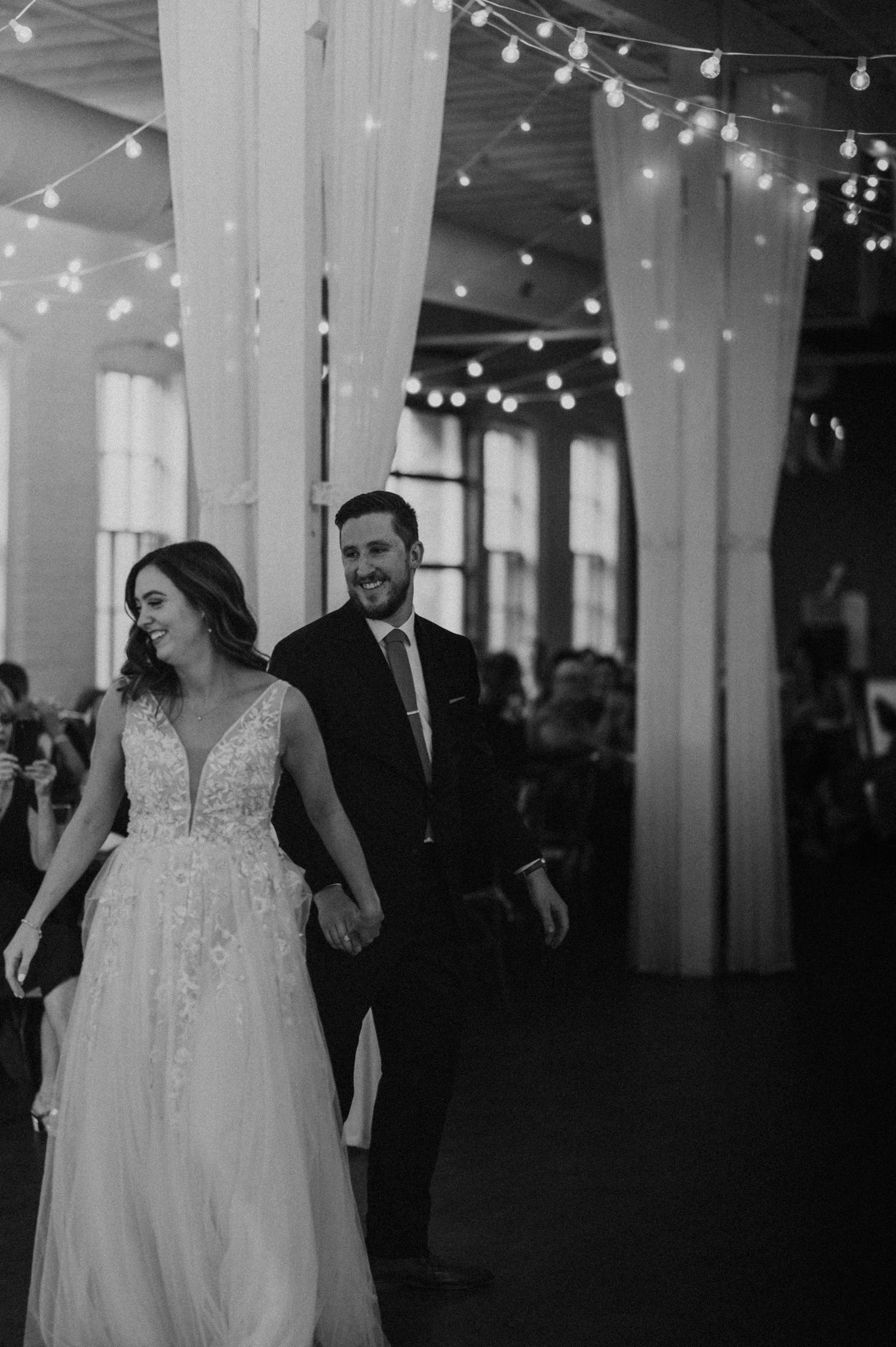 Bride and groom holding hands entering wedding reception with string lights overhead.