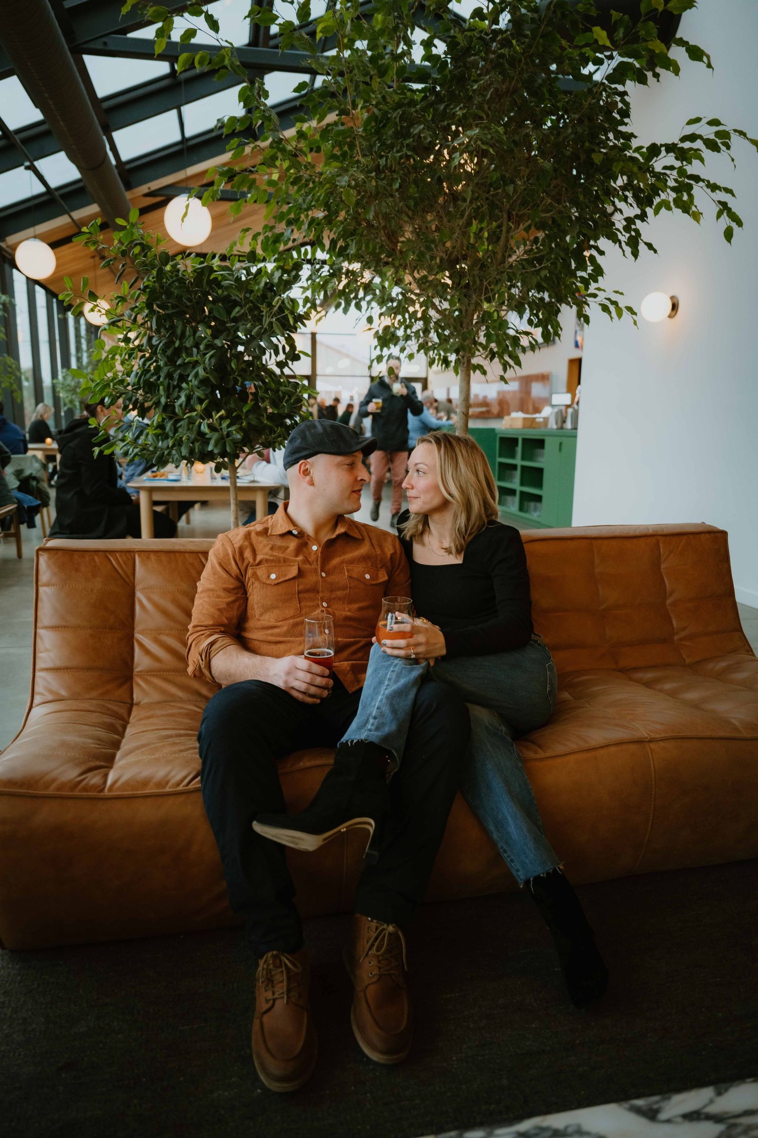 Couple sitting close on tan leather sofa sharing drinks in bright plant-filled brewery lounge.