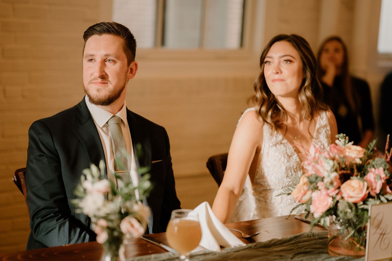 Bride and groom seated at reception table with pink floral centerpiece during wedding speeches.