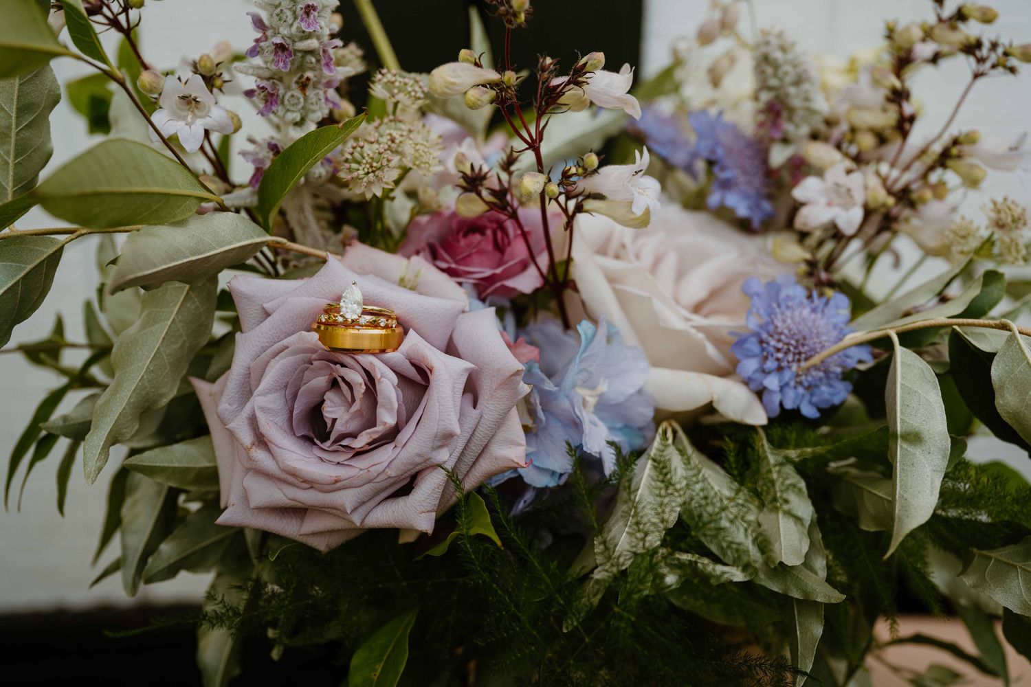 Gold wedding rings rest on a lush bouquet of mauve roses, blue hydrangeas, and white blooms.