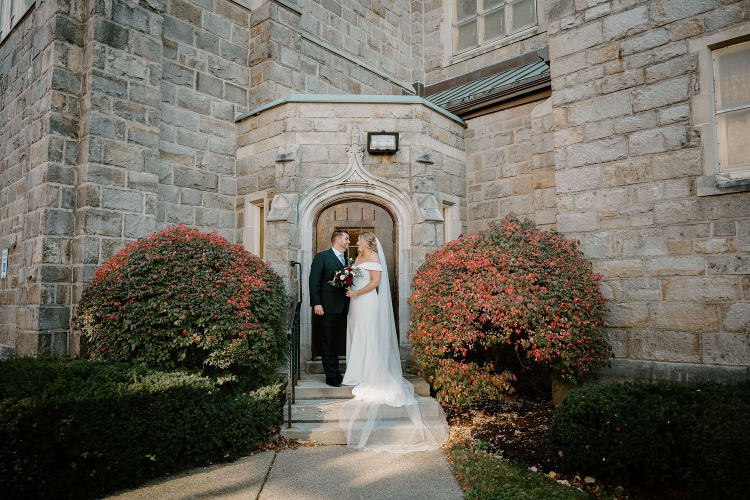 Bride and groom share romantic moment at arched stone church entrance surrounded by autumn foliage.