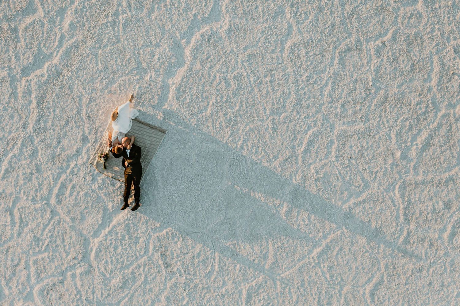 Aerial view of couple lying on white salt flats casting long dramatic shadows.