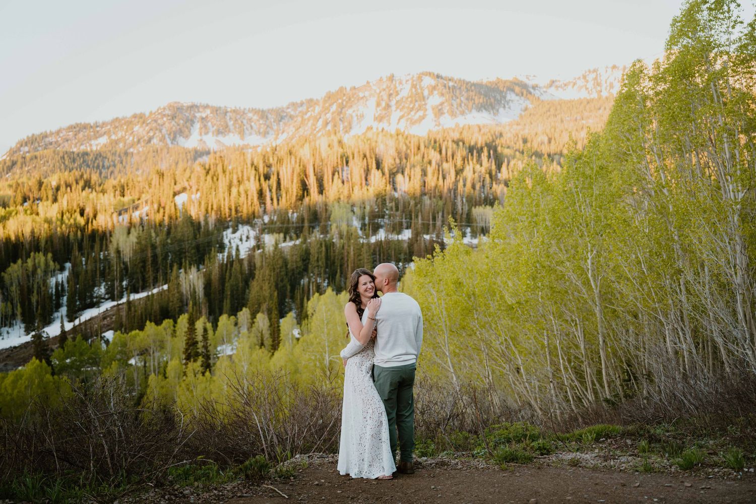 Bride and groom embrace on mountain trail surrounded by spring aspens and snow-capped peaks.