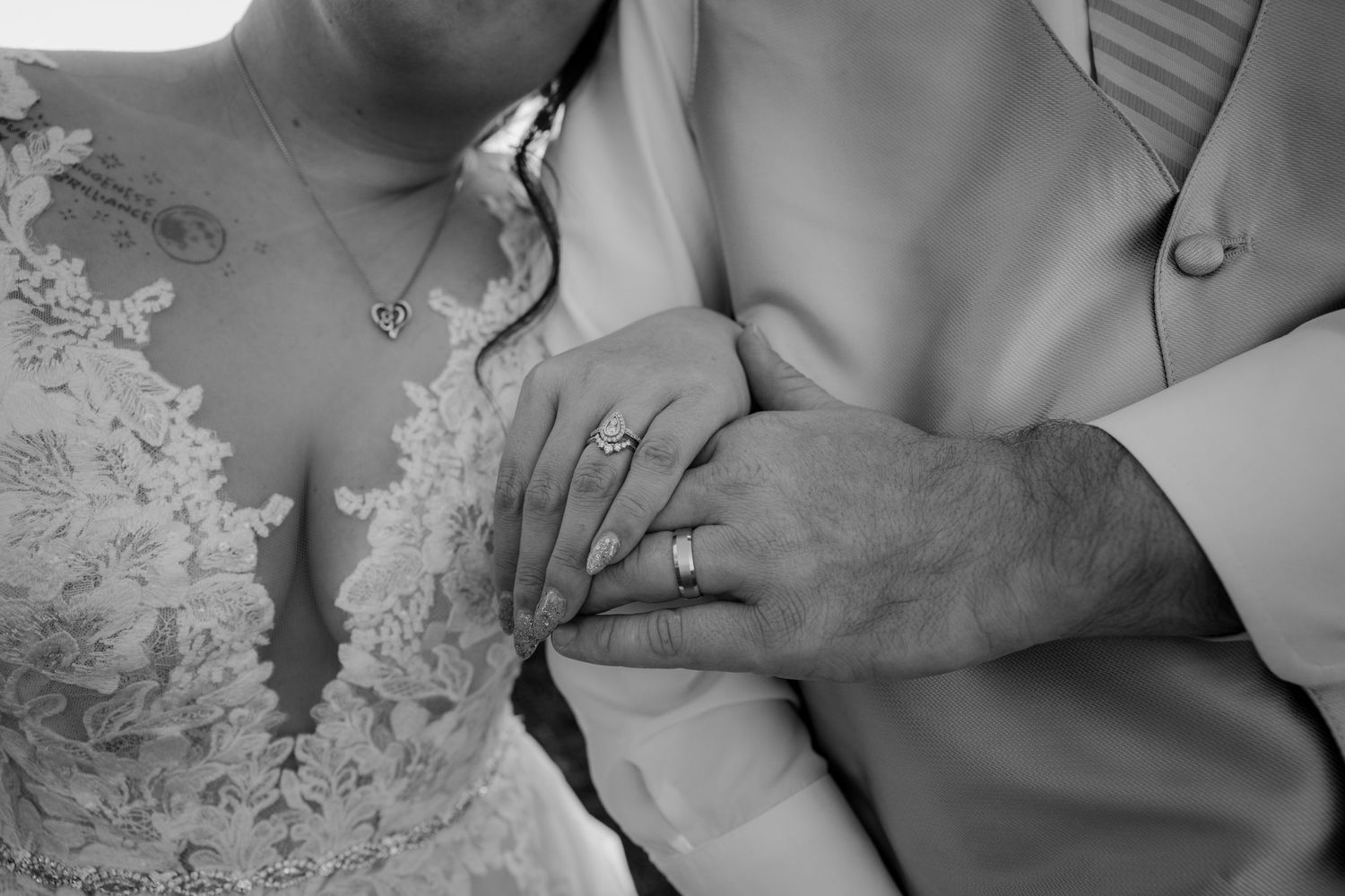 Black and white close-up of bride and groom's hands showing wedding rings against lace bridal gown.