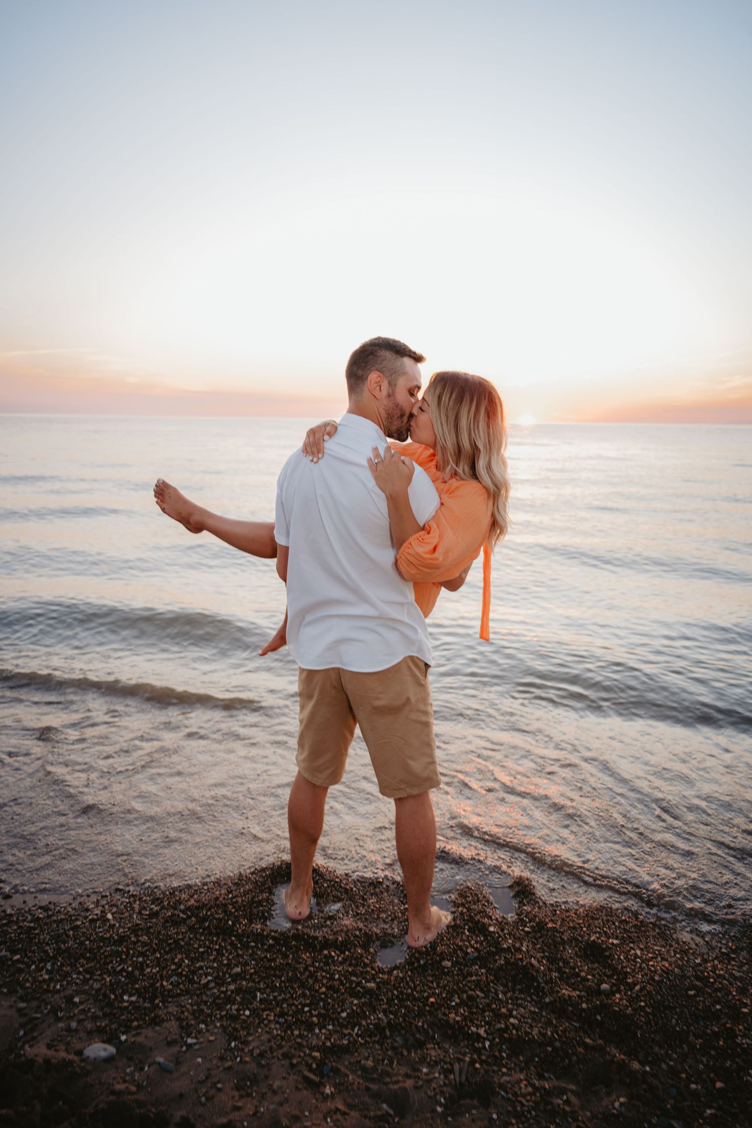 Couple sharing a romantic kiss at sunset on a rocky beach, man lifting woman in orange dress.