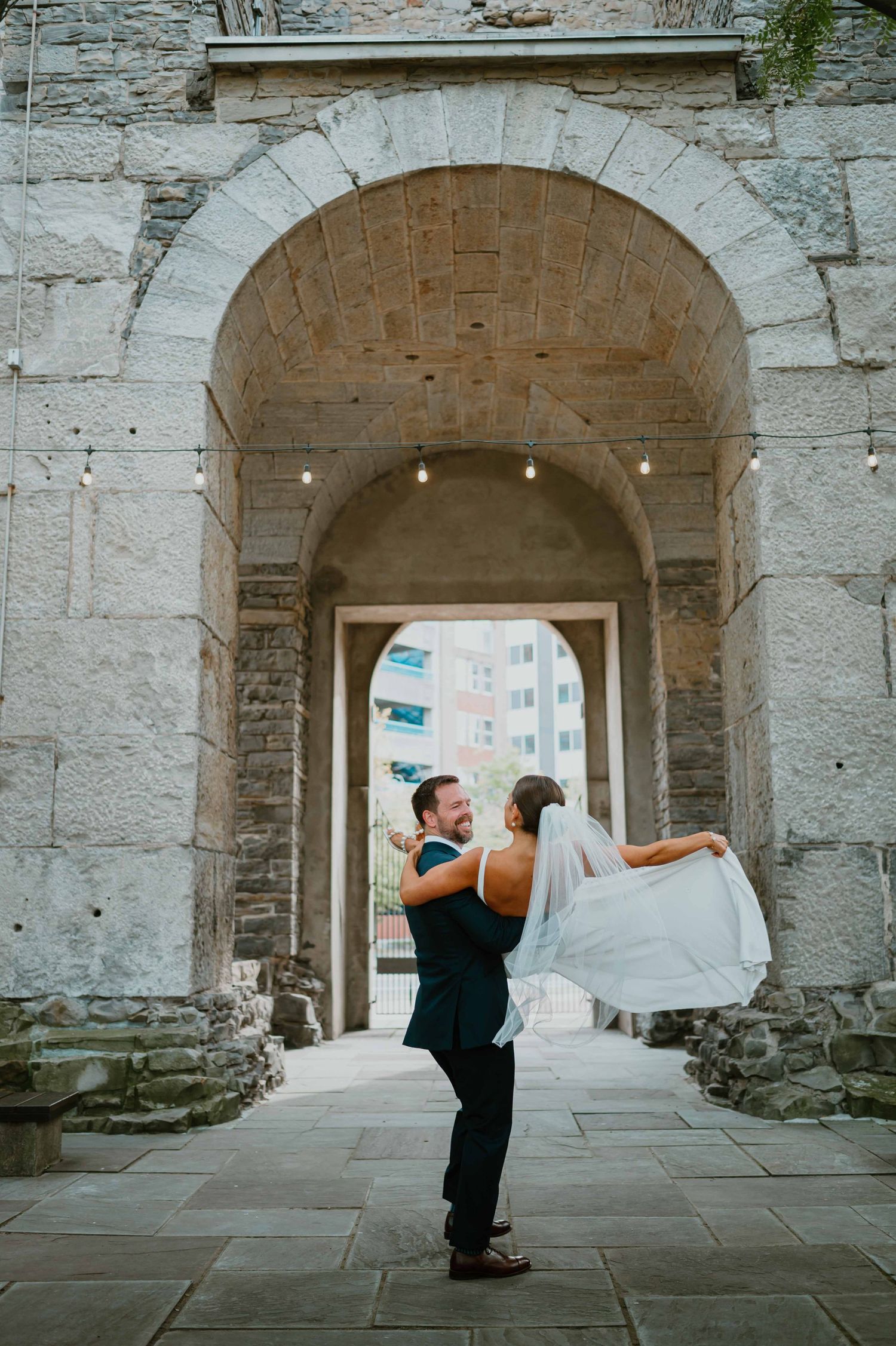 Groom lifts laughing bride as her veil flows beneath a rustic stone arch with bistro lights at wedding venue.