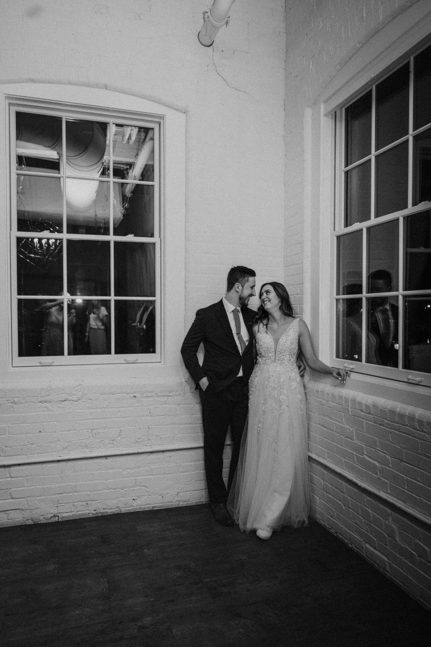 Black and white photo of bride and groom leaning together by large windows in brick venue.