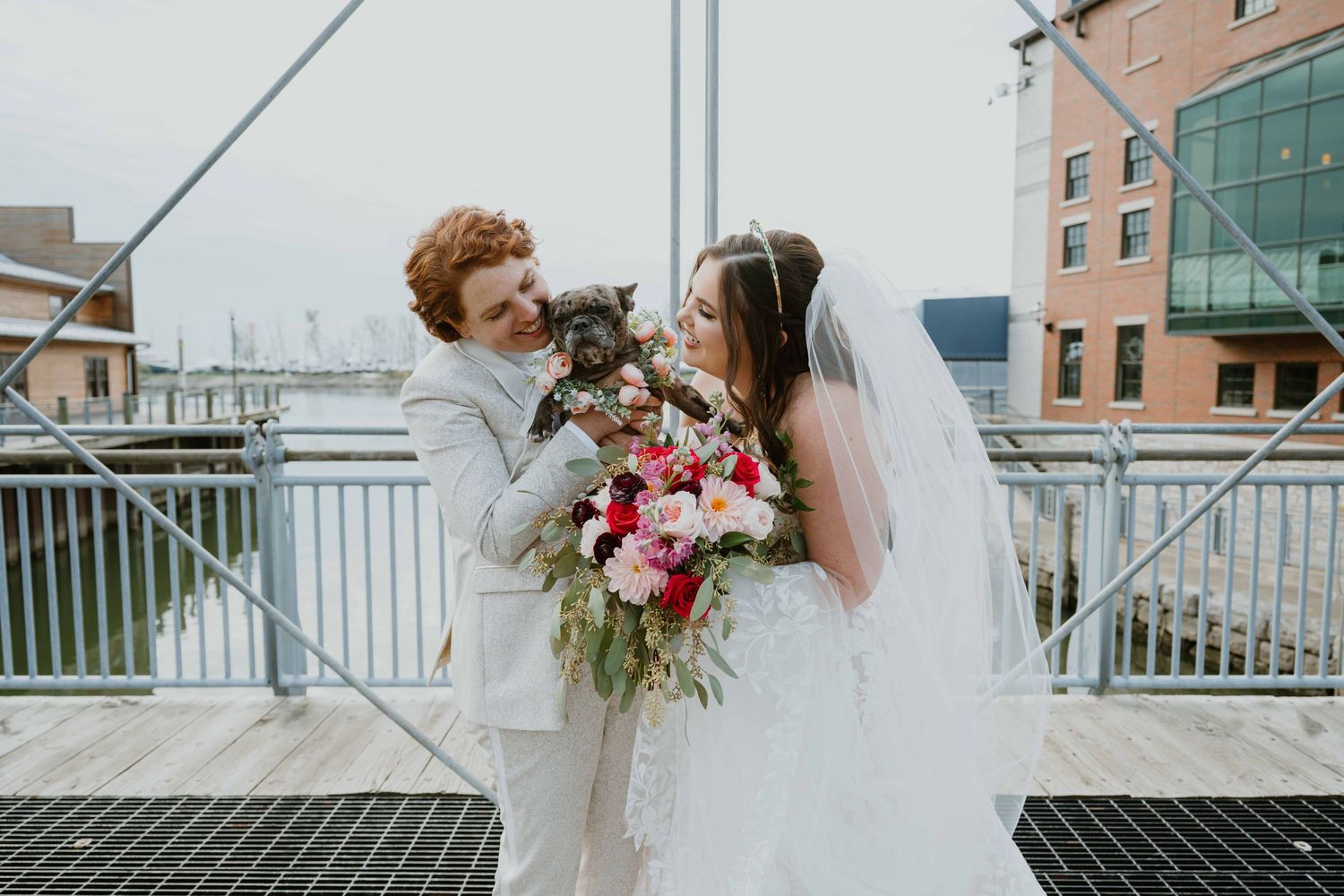 Newlyweds hold small dog while bride carries vibrant floral bouquet on outdoor bridge.