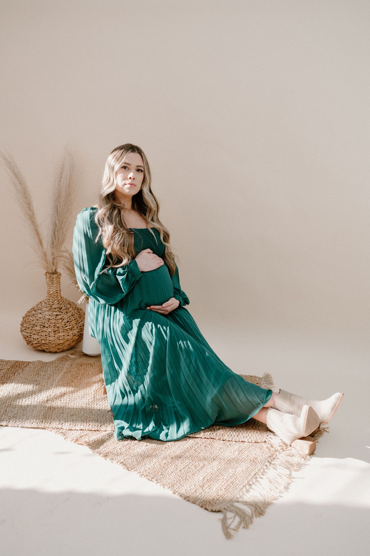 Pregnant woman in teal green maxi dress sitting on jute rug holding baby bump in bright boho studio.