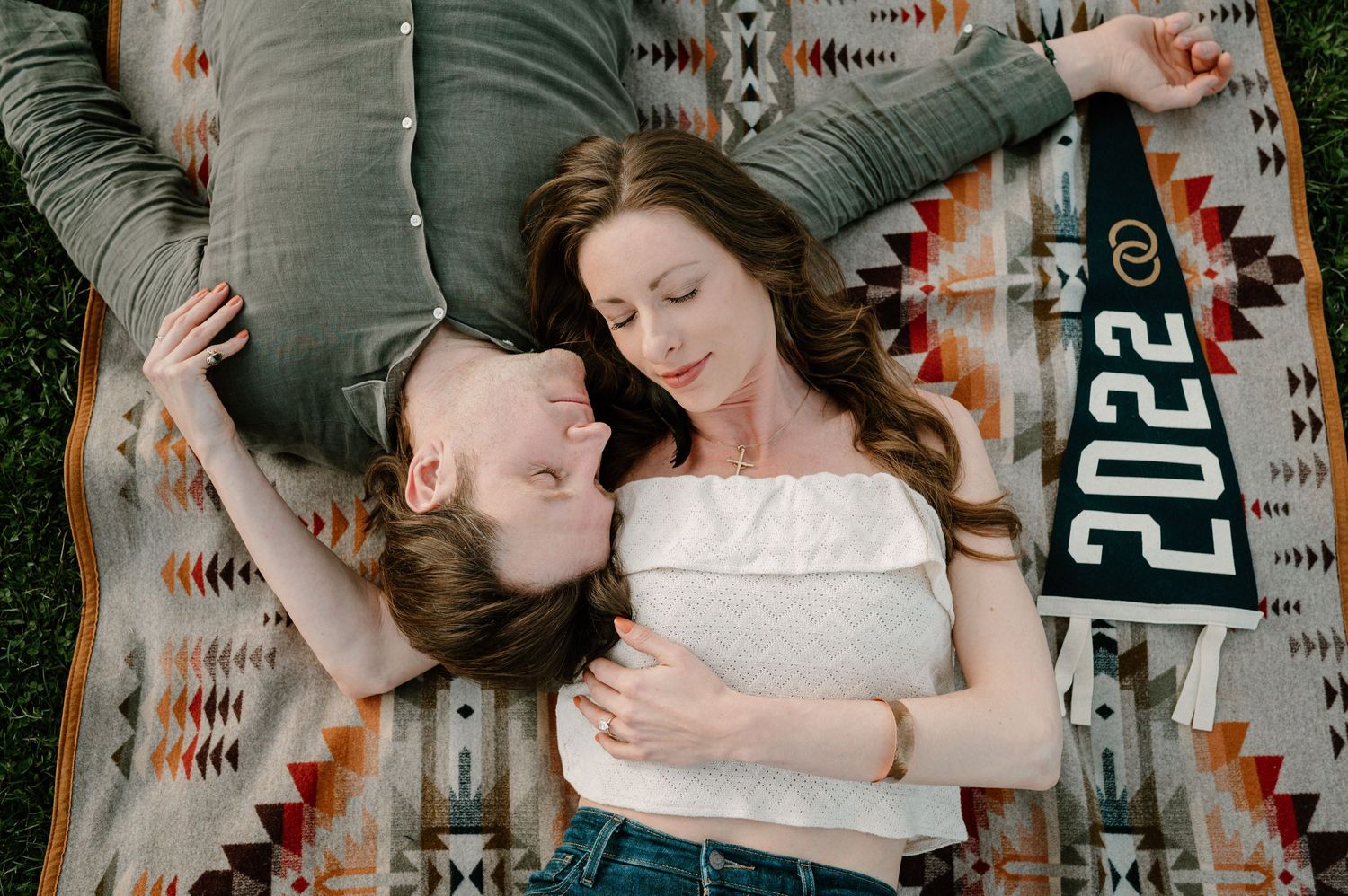 Couple lies on a Pendleton blanket outdoors holding a 2022 pennant during their engagement photo session.