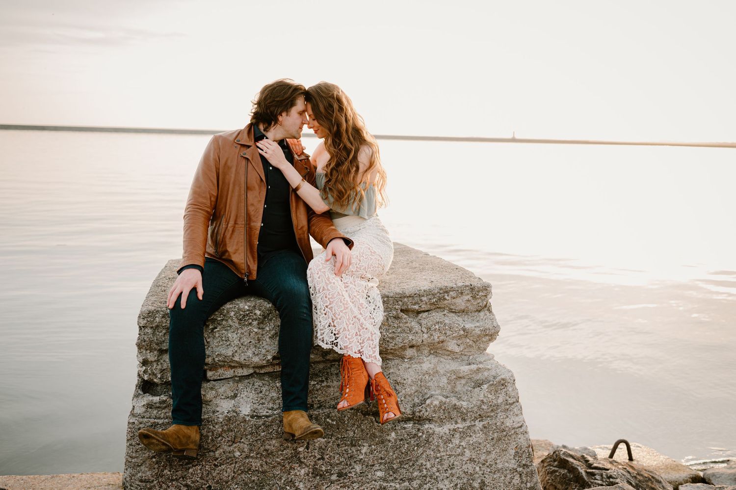 Couple shares a kiss while seated on lakeside rocks during a romantic golden hour engagement session.