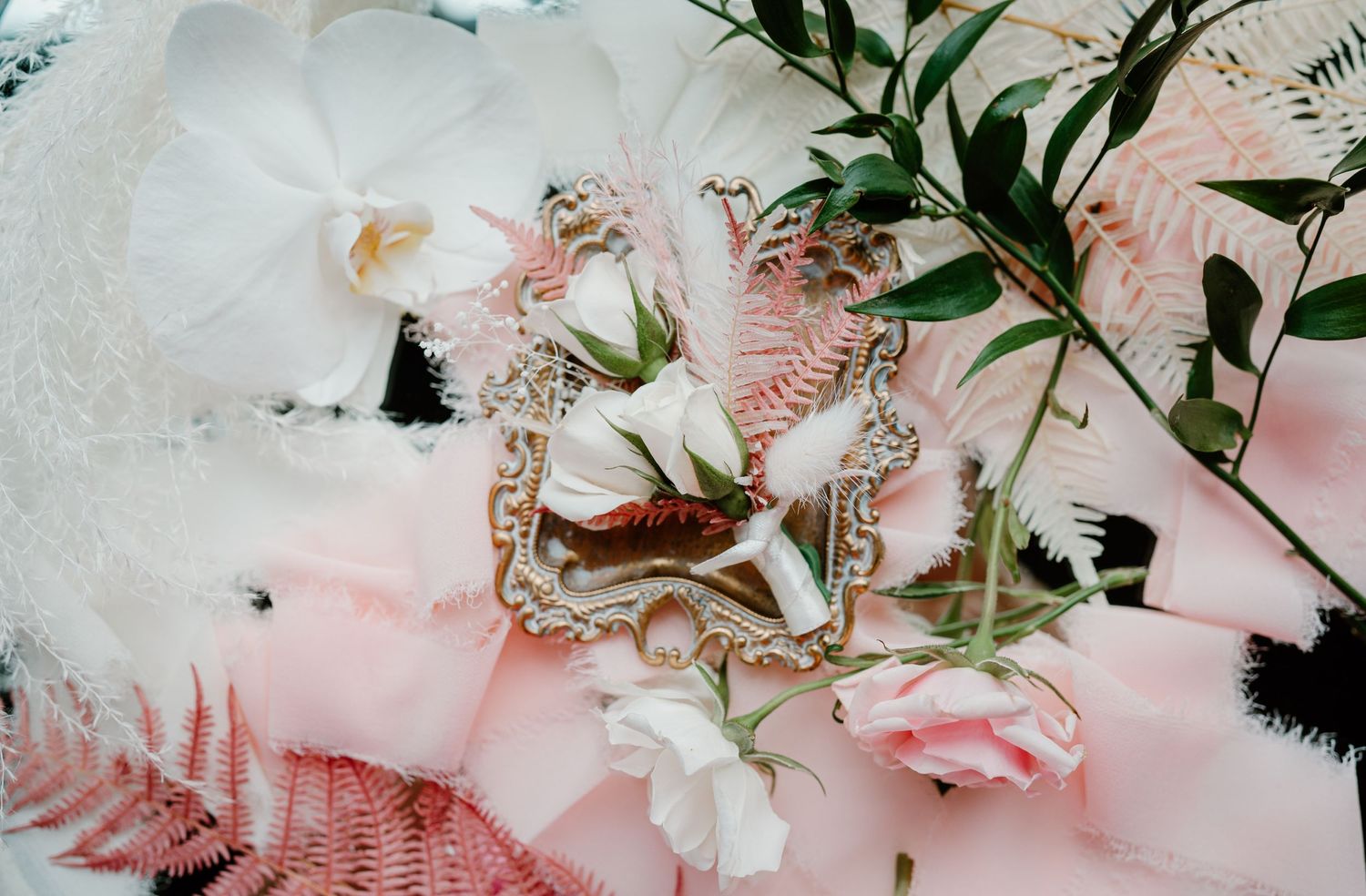 Romantic bridal detail flat lay with white roses, orchids, greenery, and ornate vintage mirror frame.