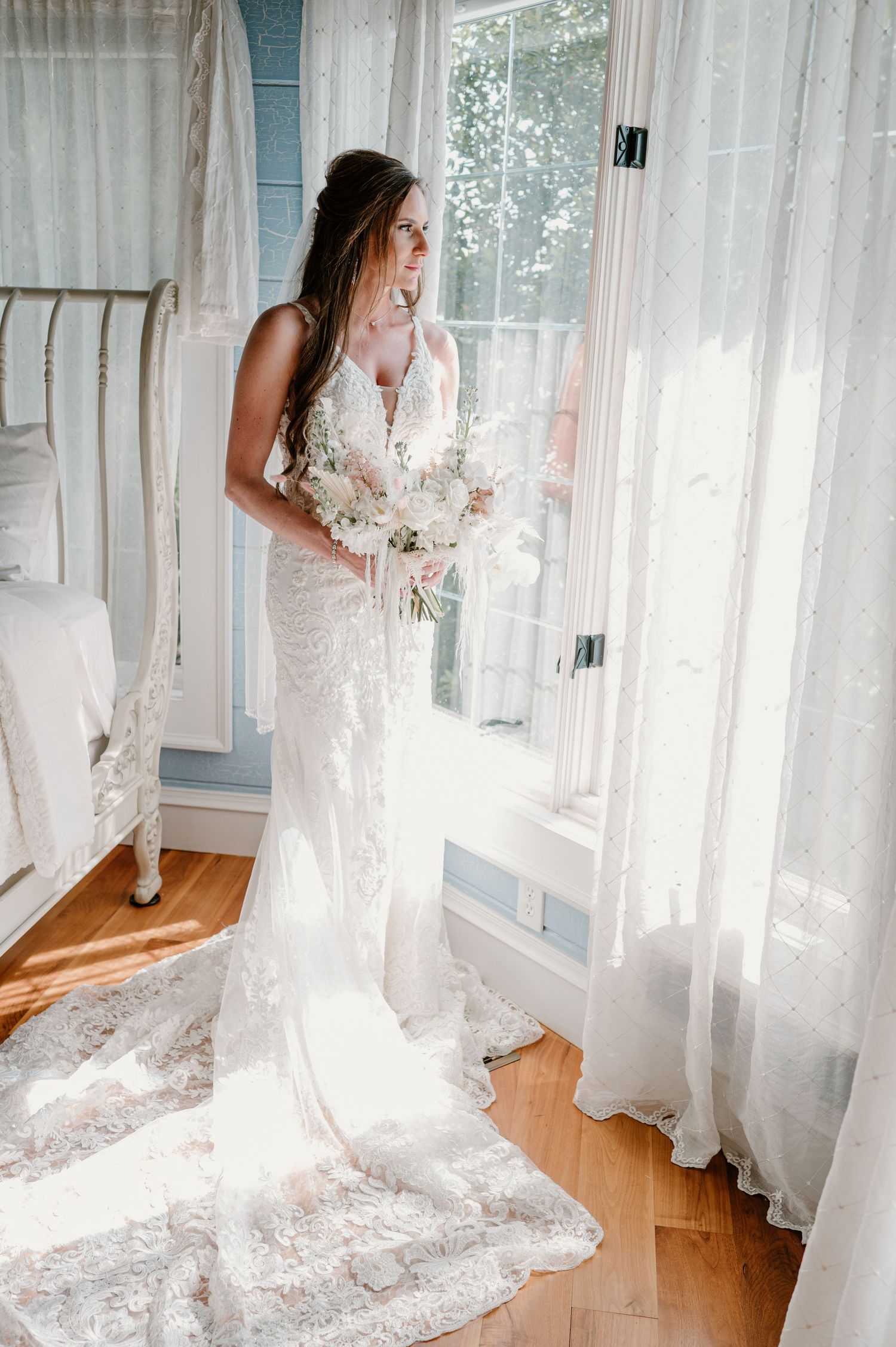 Radiant bride in lace fitted gown holds white bouquet while gazing out a sunlit window indoors.