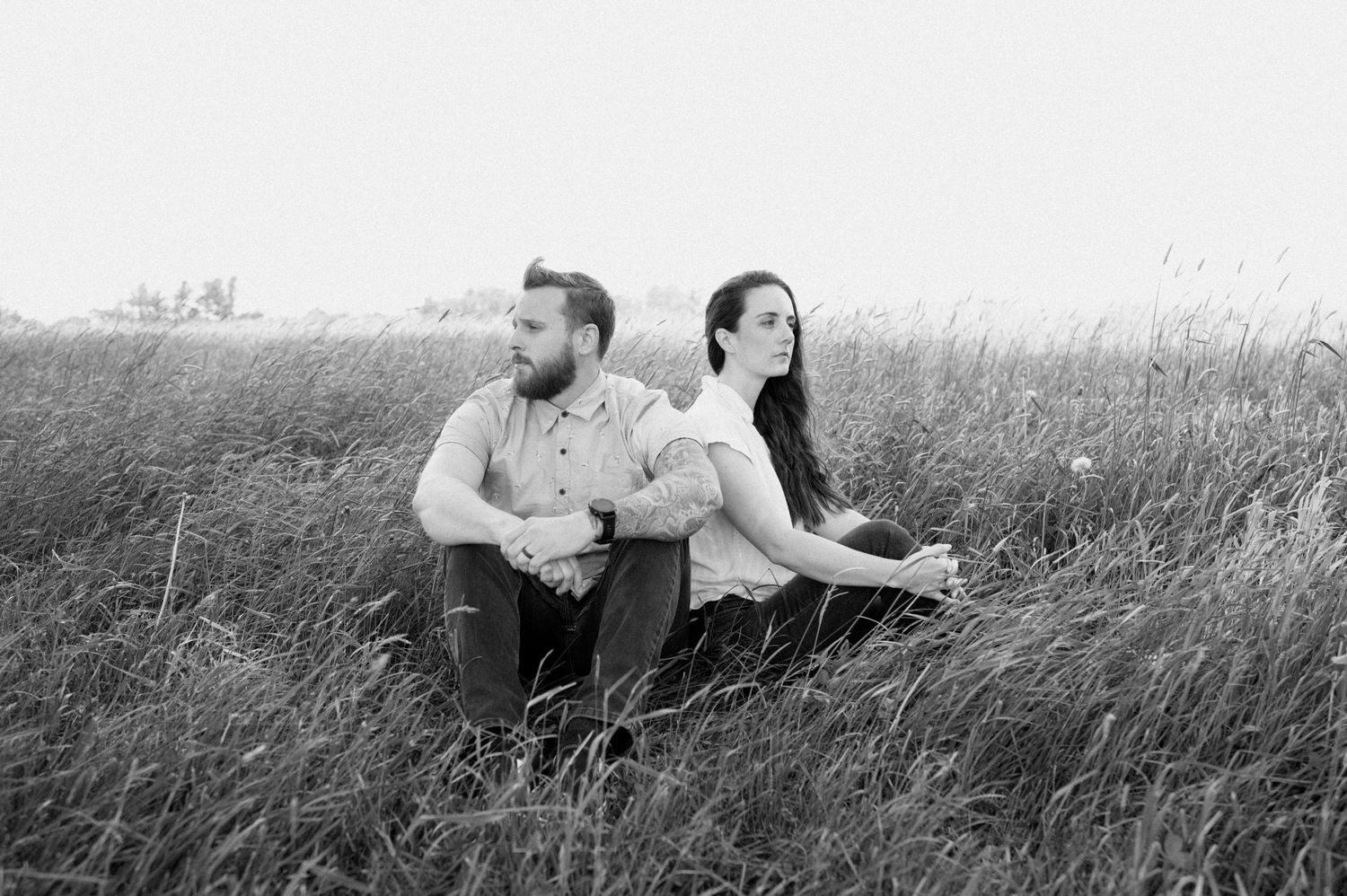 Black and white photo of couple sitting back to back in tall grass field.