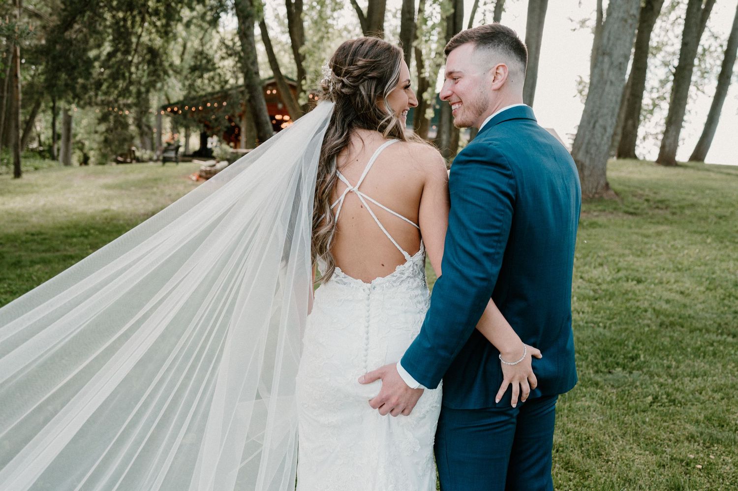 Bride and groom share a romantic moment outdoors, her lace backless gown and flowing veil catching the breeze.