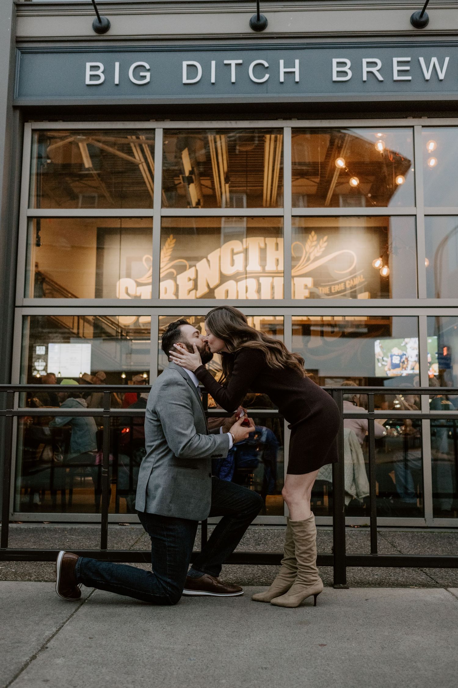 Man proposes on one knee outside Big Ditch Brewing Company as woman leans in for a kiss.