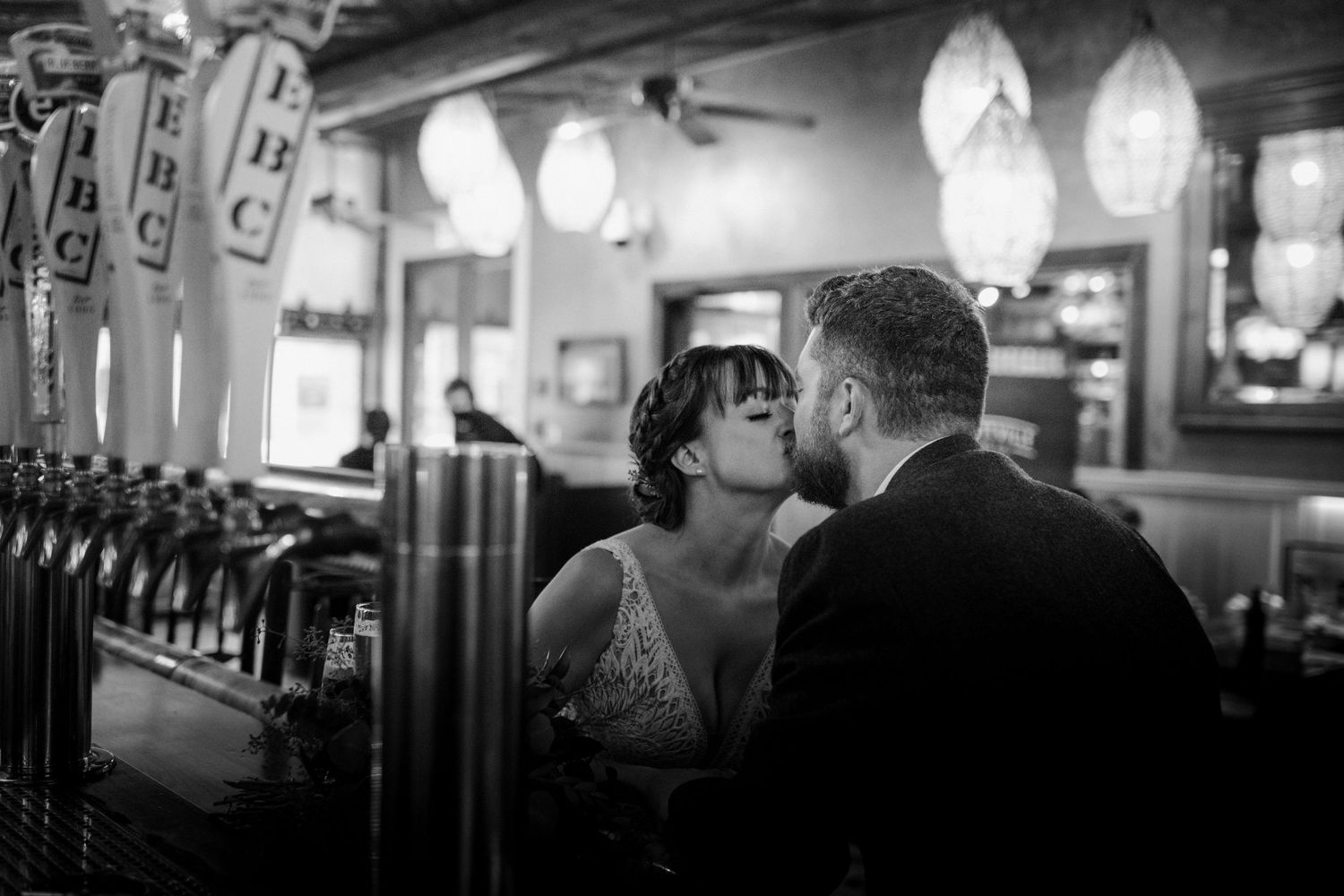 Black and white photo of bride and groom sharing a kiss at a bar with beer taps in the foreground.
