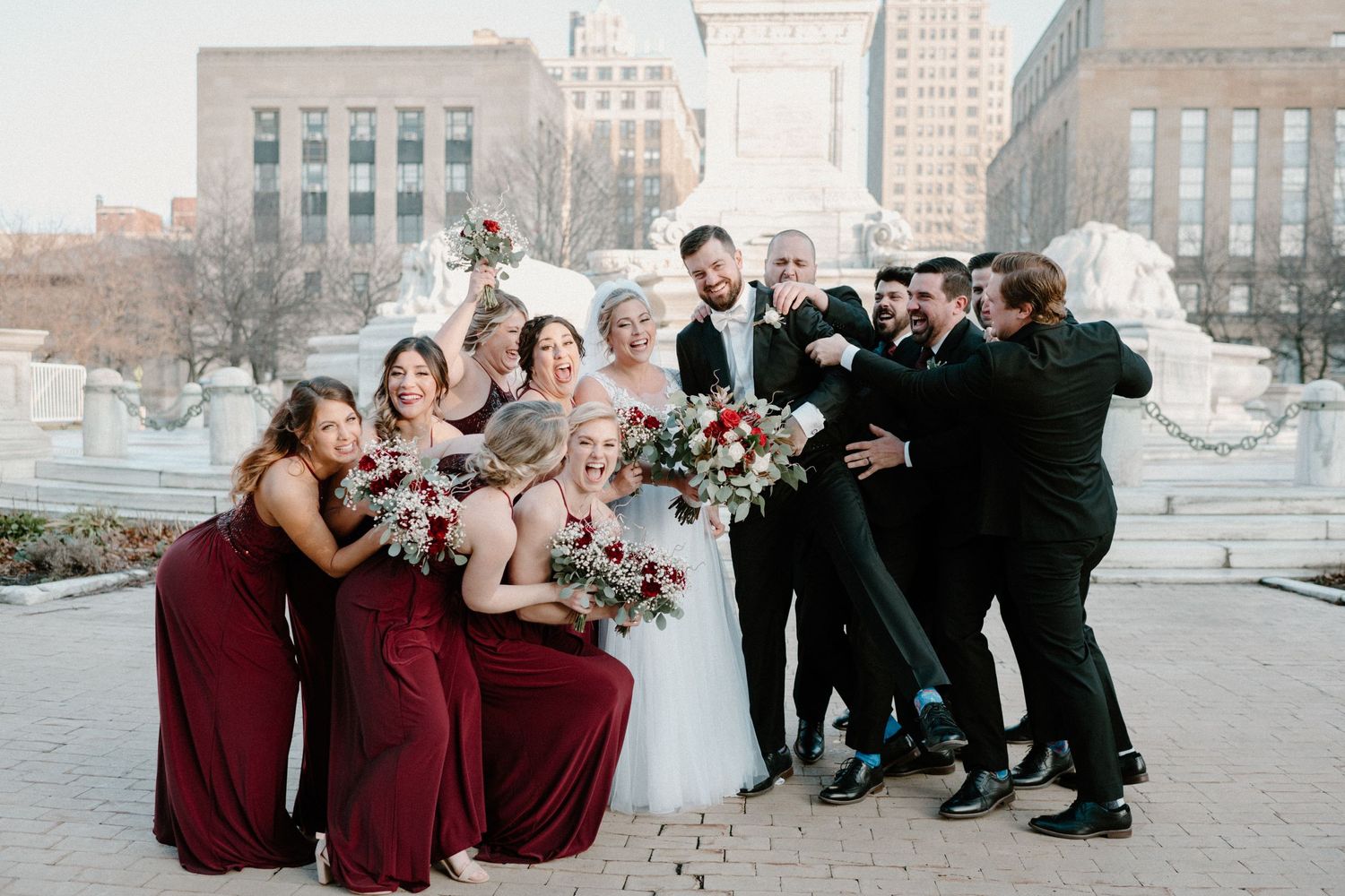 Wedding party in burgundy and black attire celebrate joyfully in front of a fountain in the city.