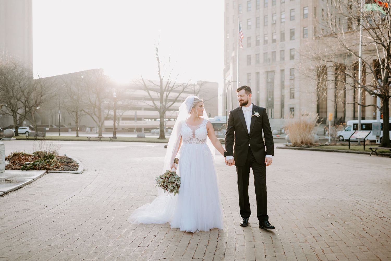Bride and groom hold hands walking through an urban plaza in golden sunlight on their wedding day.