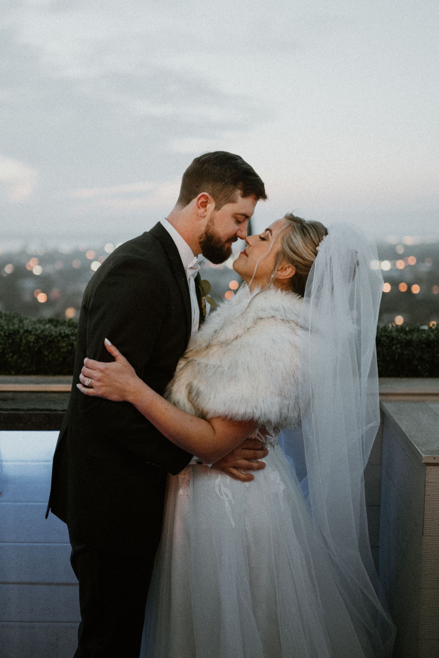 Bride and groom share a tender kiss on a rooftop at dusk, bride wearing a fur stole and veil.