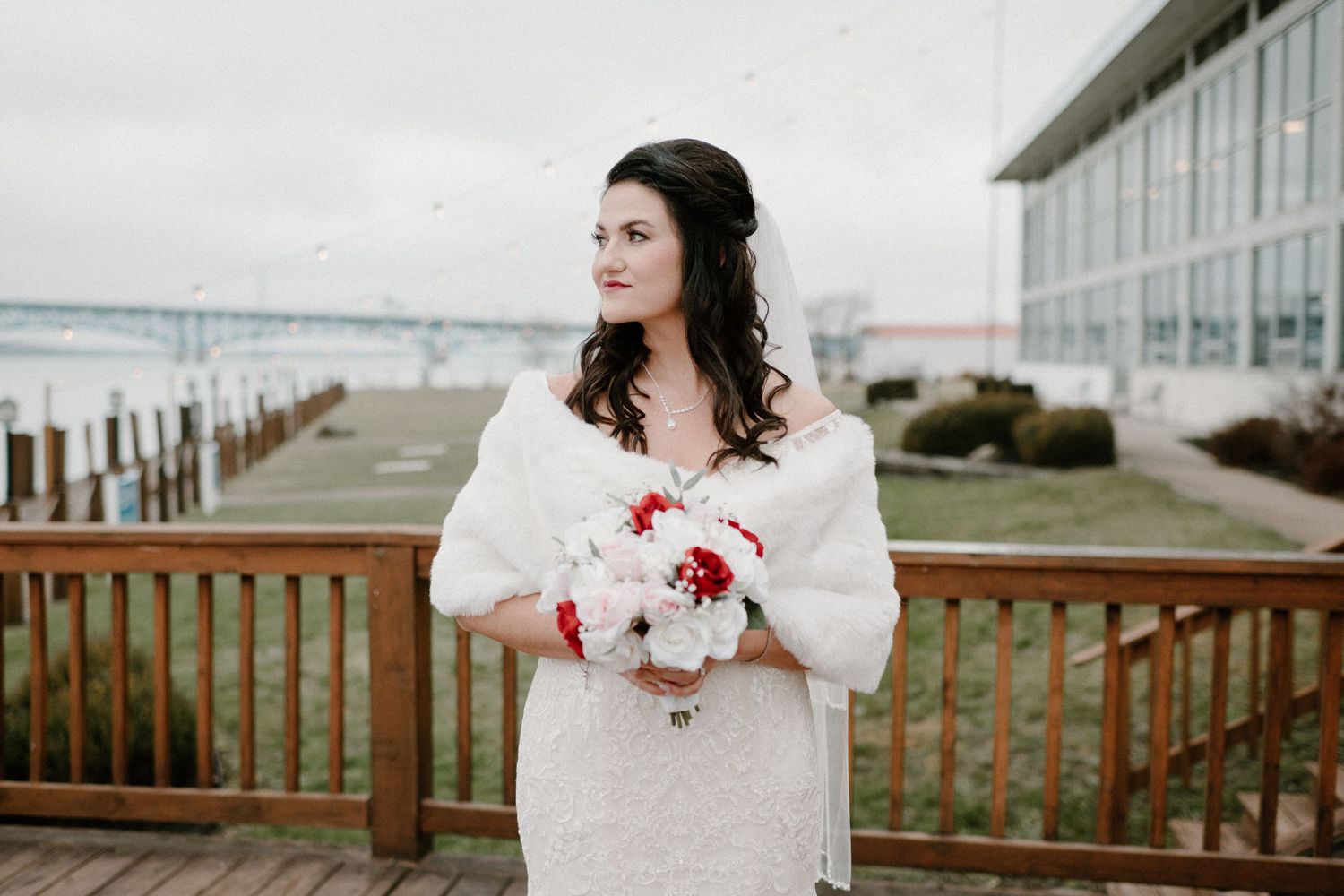 Winter bride in white lace gown and faux fur wrap holds red and white bouquet on waterfront deck.