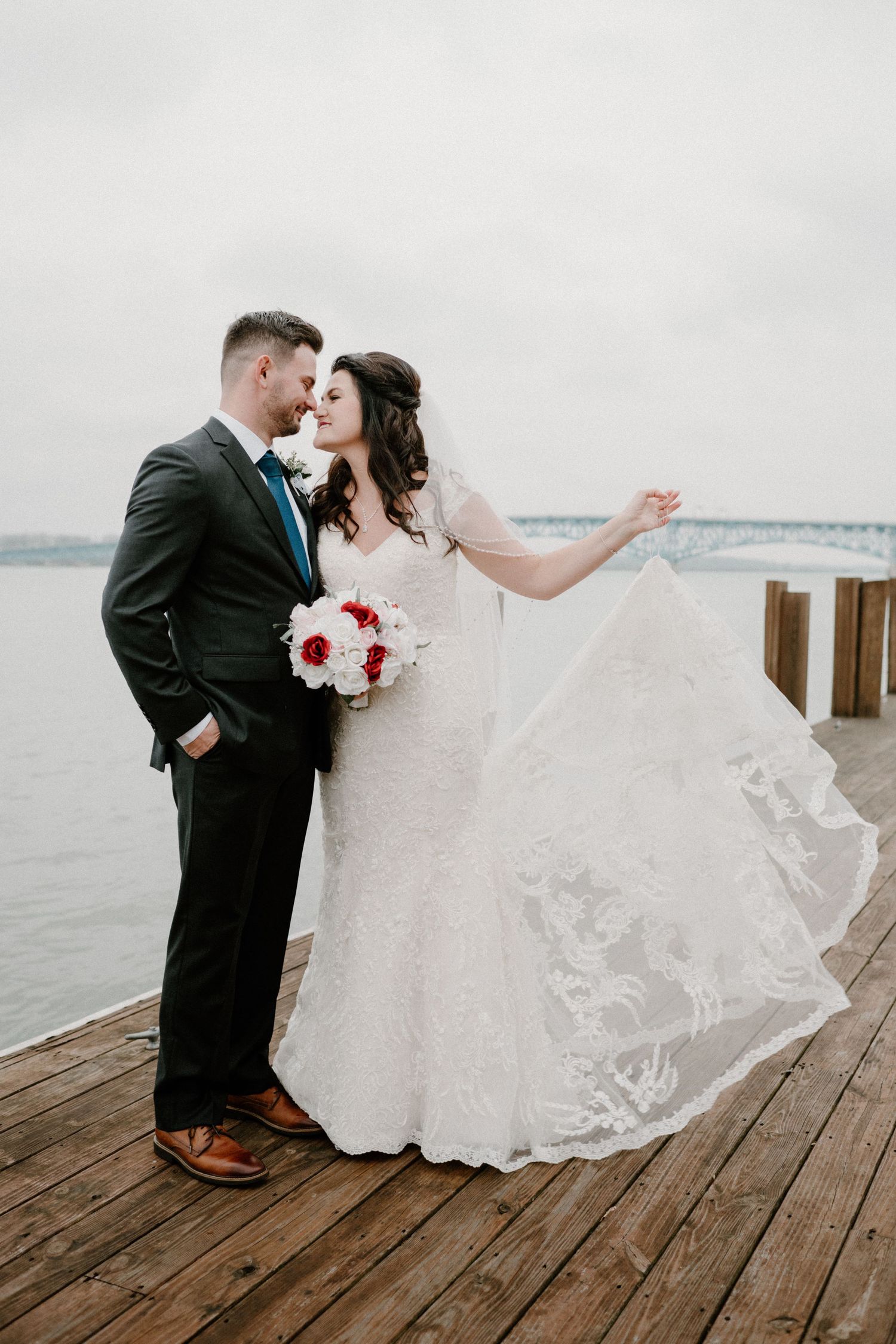 Bride and groom share a tender moment on a wooden dock with lace gown flowing in the waterfront breeze.