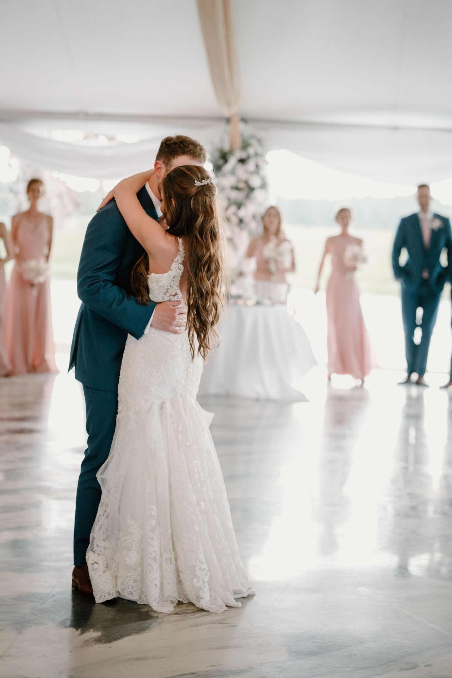 Bride and groom share their first dance under a white tent while wedding party looks on in soft focus.
