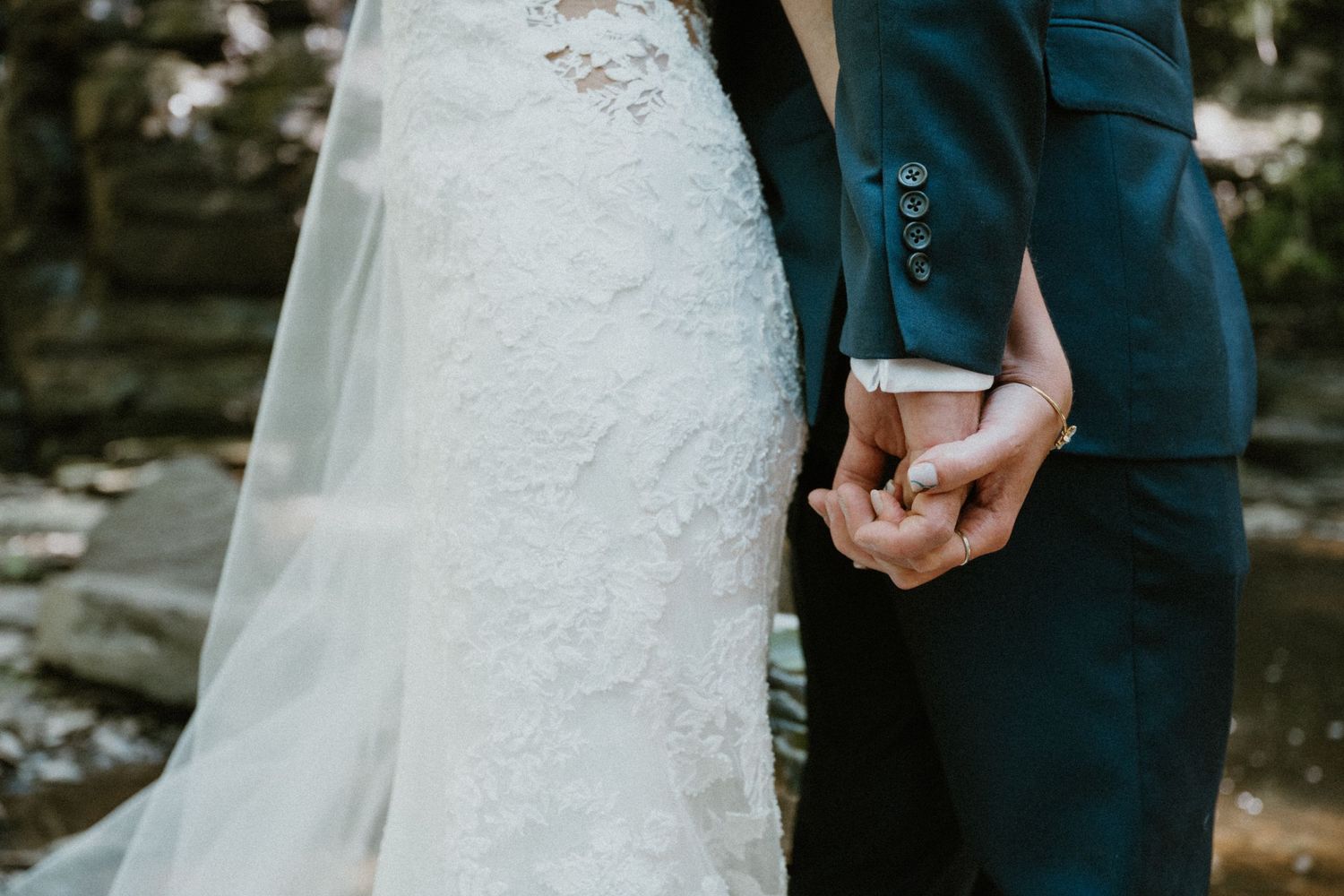 Close-up wedding photo of bride and groom holding hands, showcasing lace gown and navy blue suit details.