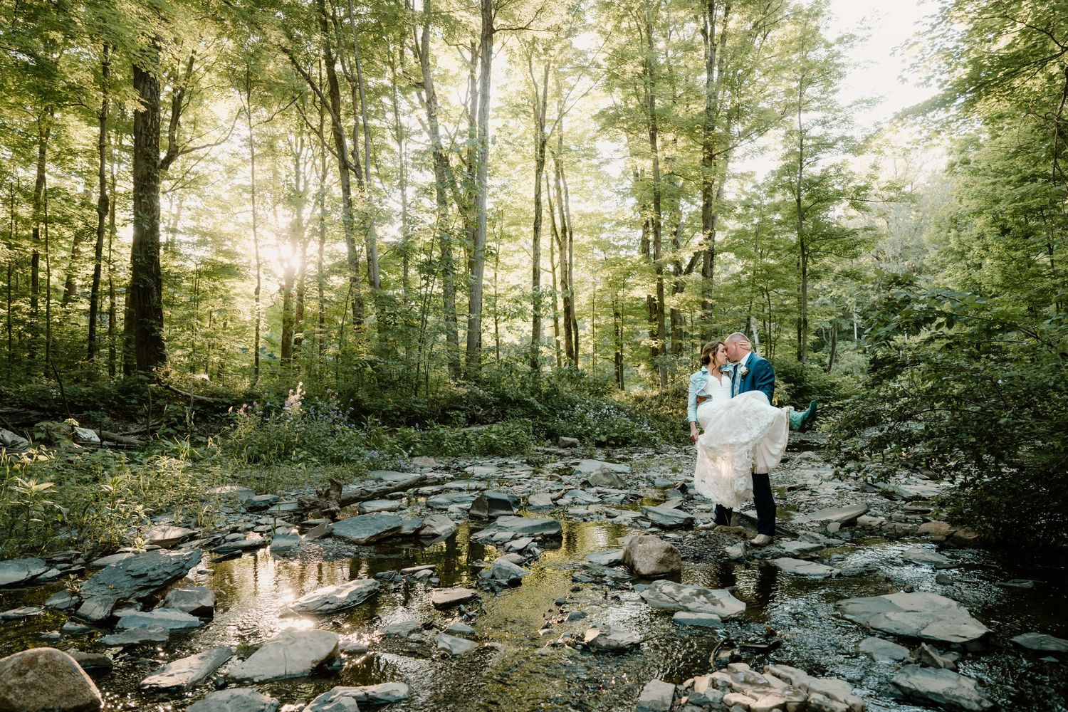 Romantic wedding photo of groom carrying bride through a sunlit forest creek surrounded by lush green trees.